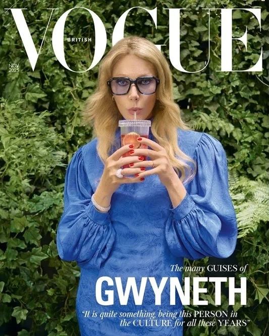 Eine Frau mit blonden, lockigen Haaren, trägt eine blaue Bluse und große Brillengläser, trinkt einen pinken Drink aus einem transparenten Becher mit Strohhalm, im Hintergrund grüne Blätter auf einem Cover des Vogue Magazins November 2025.