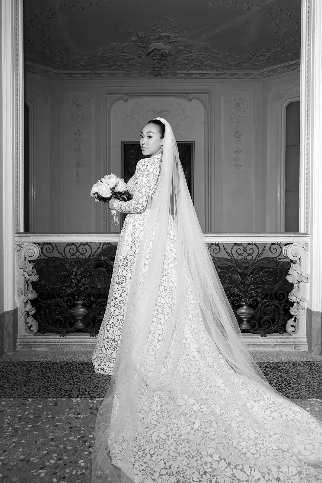 Schwarzweiß-Foto einer Braut in Hochzeitskleid mit Blumenstrauß in einer eleganten, ornamentierten Innenraum.
