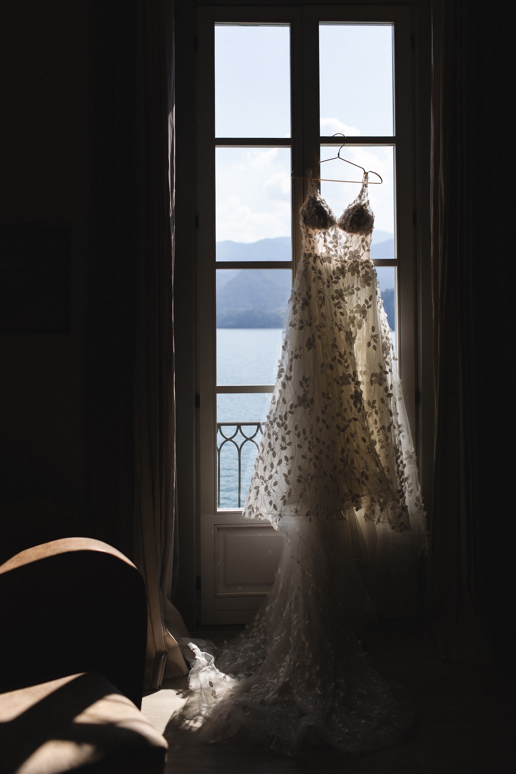 Ein Brautkleid hängt an einem Kleiderbügel vor einem Fenster mit Blick auf Berge und ein Gewässer, Sonnenlicht fällt auf den Saum des Kleids.