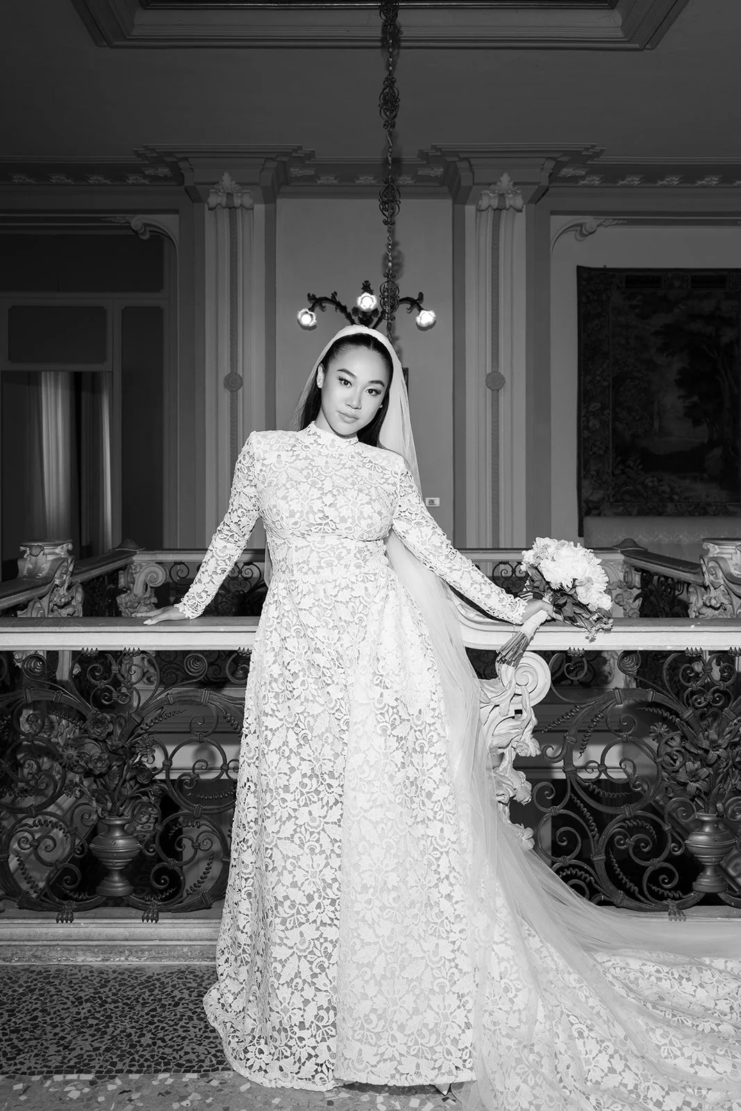 Schwarz-Weiß-Fotografie einer jungen Frau im Hochzeitskleid mit Blumenstrauß, stehend an einer Balustrade in einem eleganten, historischen Raum mit Kronleuchter und Wanddekoration.