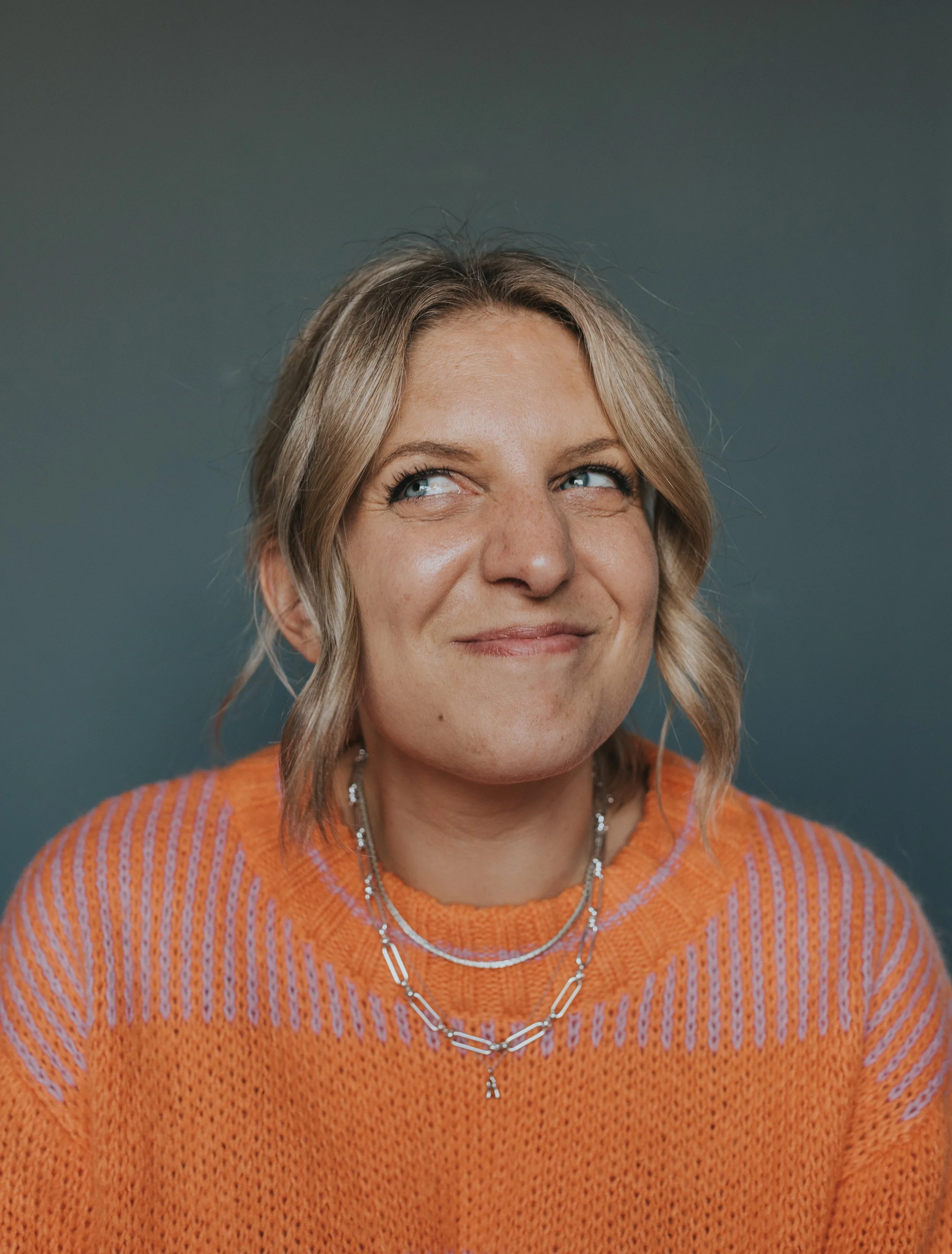 Close-up of a smiling woman with blonde hair styled in loose waves, wearing an orange sweater with purple stripes, layered necklaces, and a gray background.