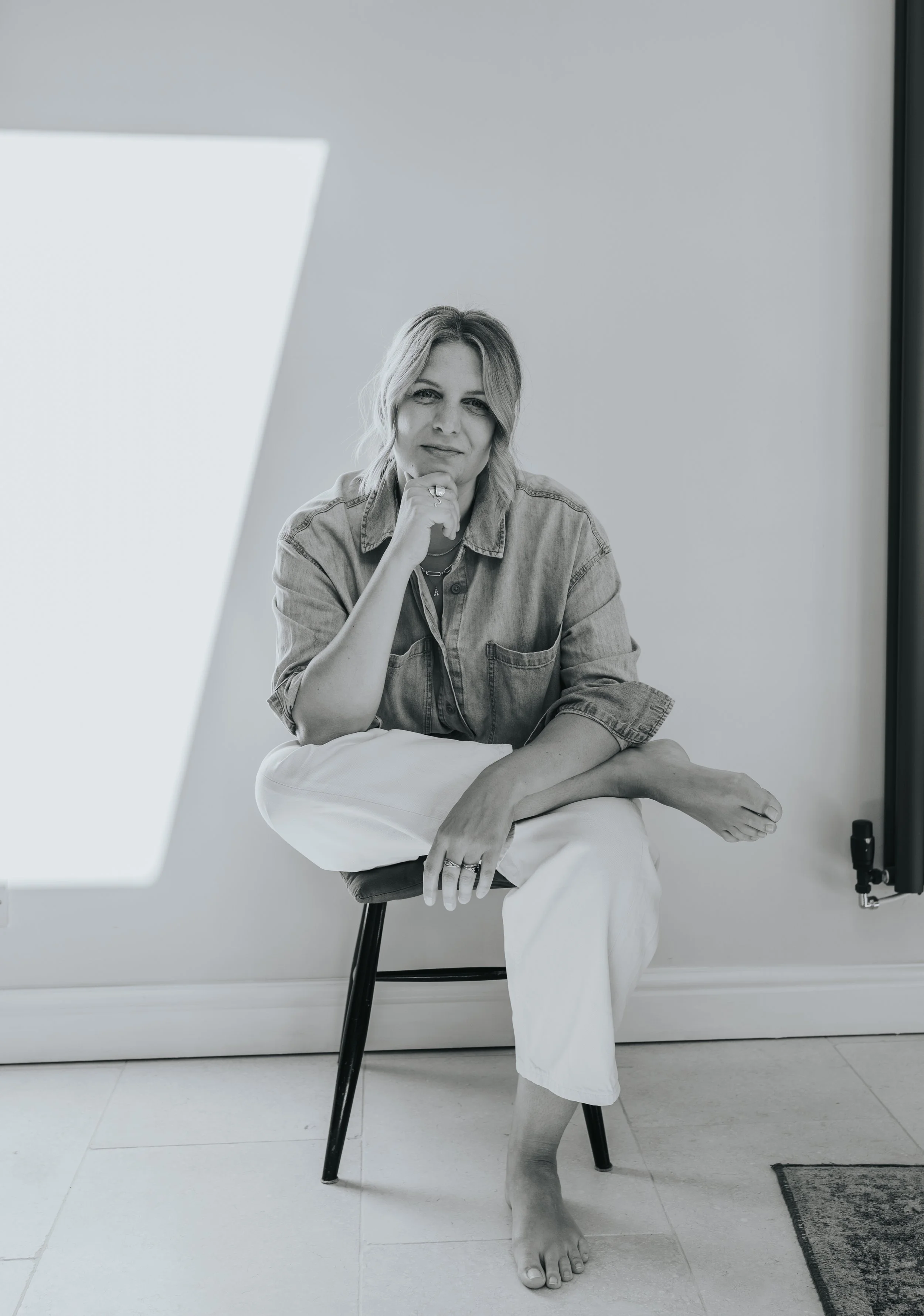 Black and white photo of a woman sitting on a chair, resting her chin on her right hand, wearing a denim shirt and white pants, with her left leg crossed over her right knee, in a room with plain walls and a window.
