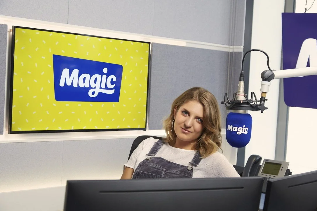 A woman sitting in a radio station studio with a large screen displaying how. The station branding features the word 'Magic' in a blue box on a yellow background with confetti.

There is a microphone with a blue cover also branded with the word 'Magic,' and radio equipment in the studio.