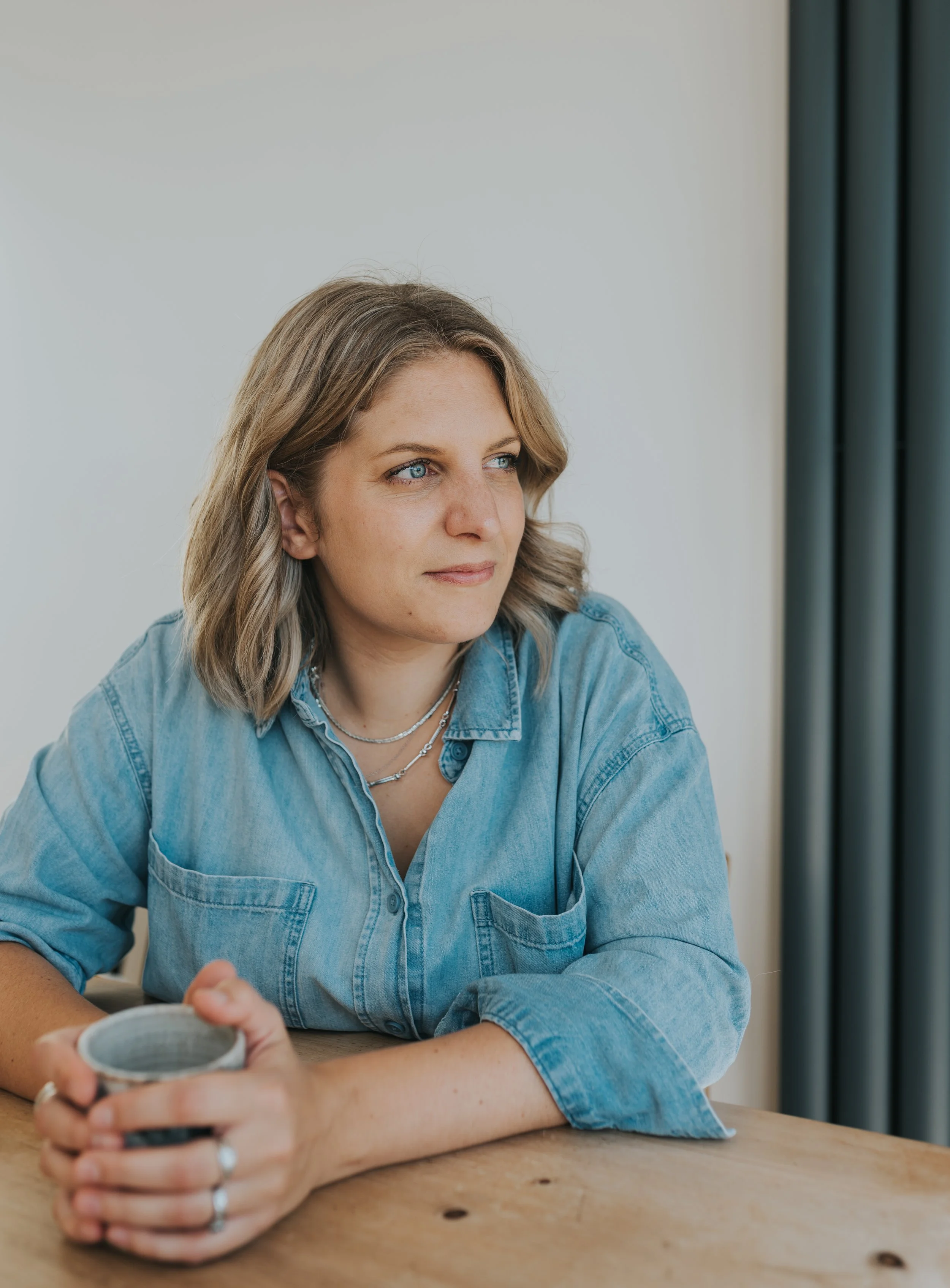 A woman with blonde hair and blue eyes sitting at a wooden table, holding a mug, wearing a denim shirt and layered necklaces, looking thoughtfully to the side near dark curtains.
