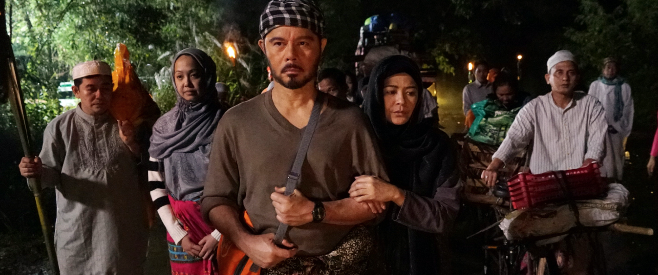 Group of people standing outdoors at night, some wearing traditional clothing and head coverings, with trees and dim lights in the background.