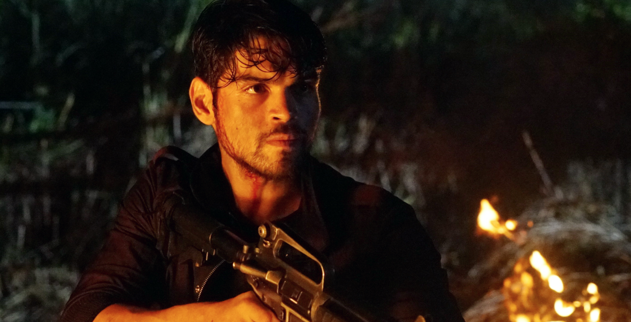 A man with dark hair and a beard holds a gun in his hand, standing outdoors at night near a small fire, with a serious expression on his face.