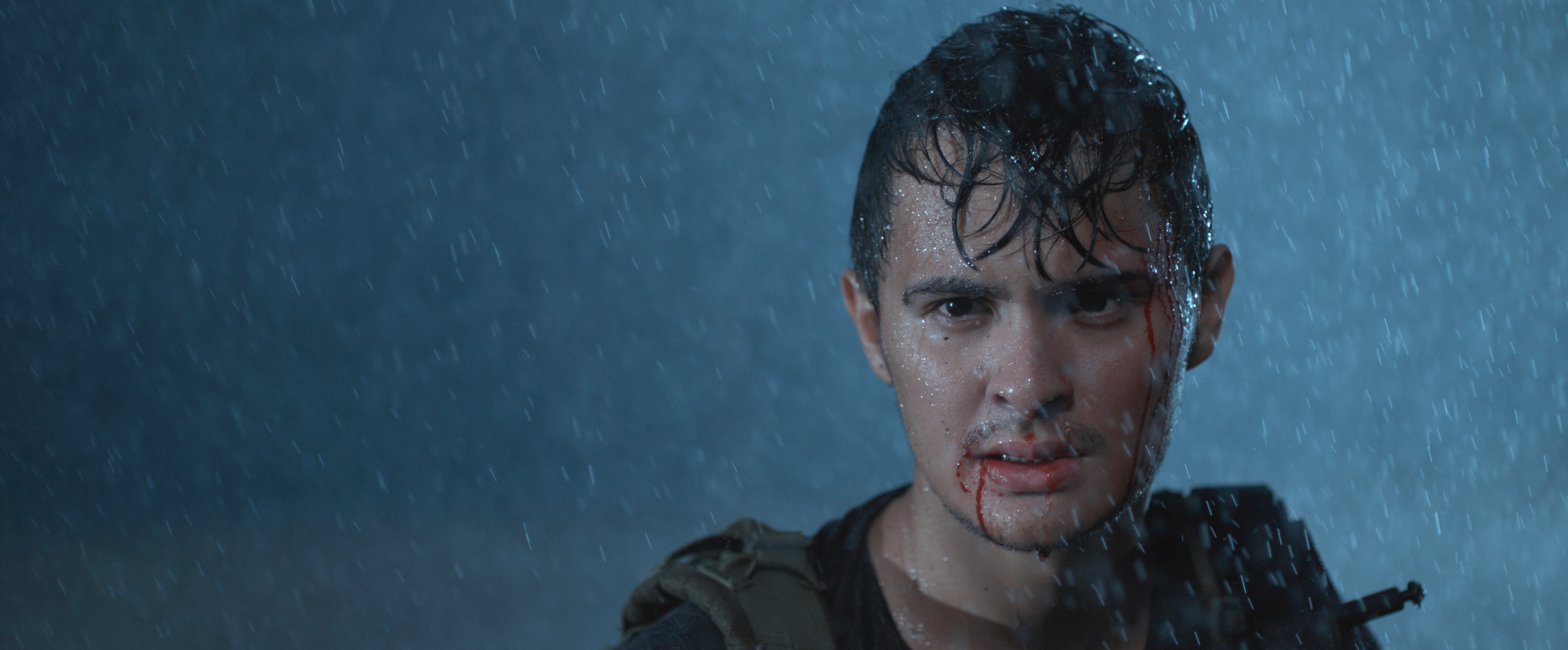 A young man with wet hair and a slight bloodstain on his face in heavy rain, looking directly at the camera with a serious expression.