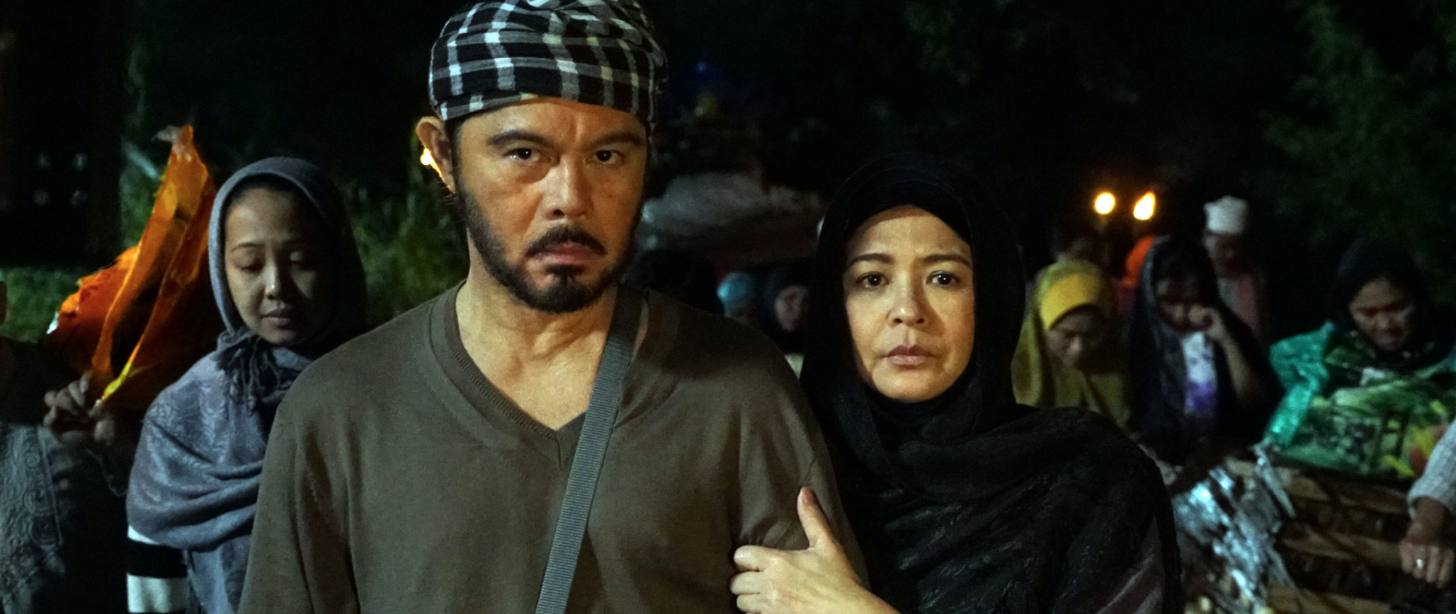 A group of people, including a man with a beard wearing a checkered cap and a woman in a black hijab, stand together at night with trees and lights in the background. They appear to be in a solemn or serious moment.
