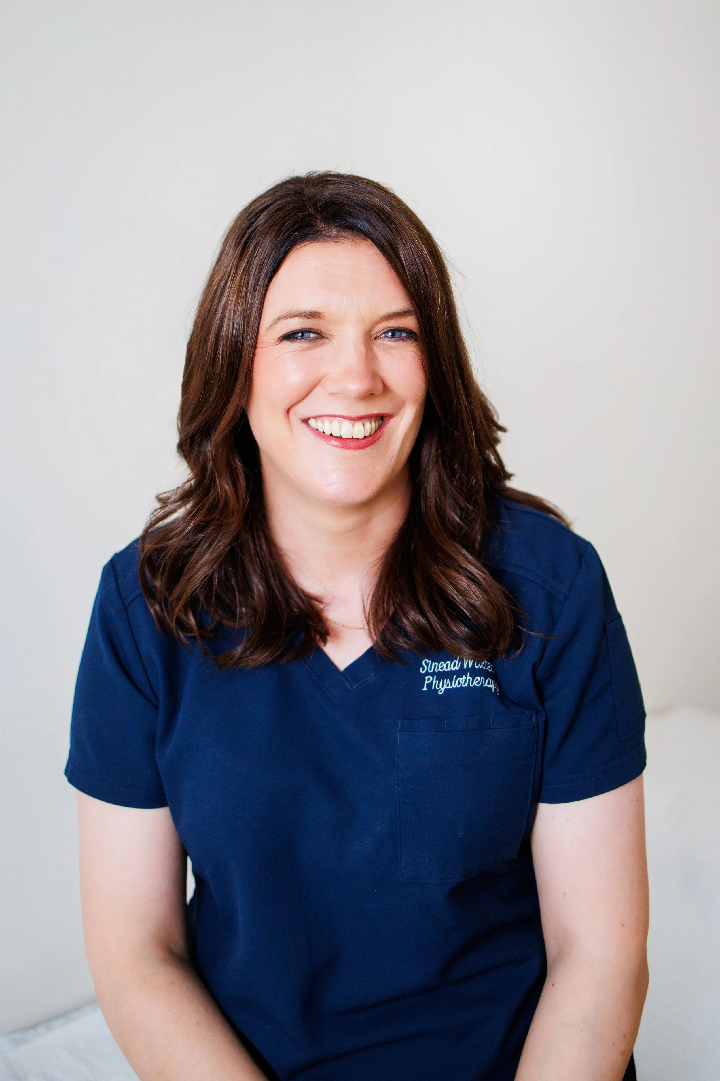 A woman with medium-length brown hair, wearing a navy blue healthcare uniform, smiling and sitting against a plain white background.
