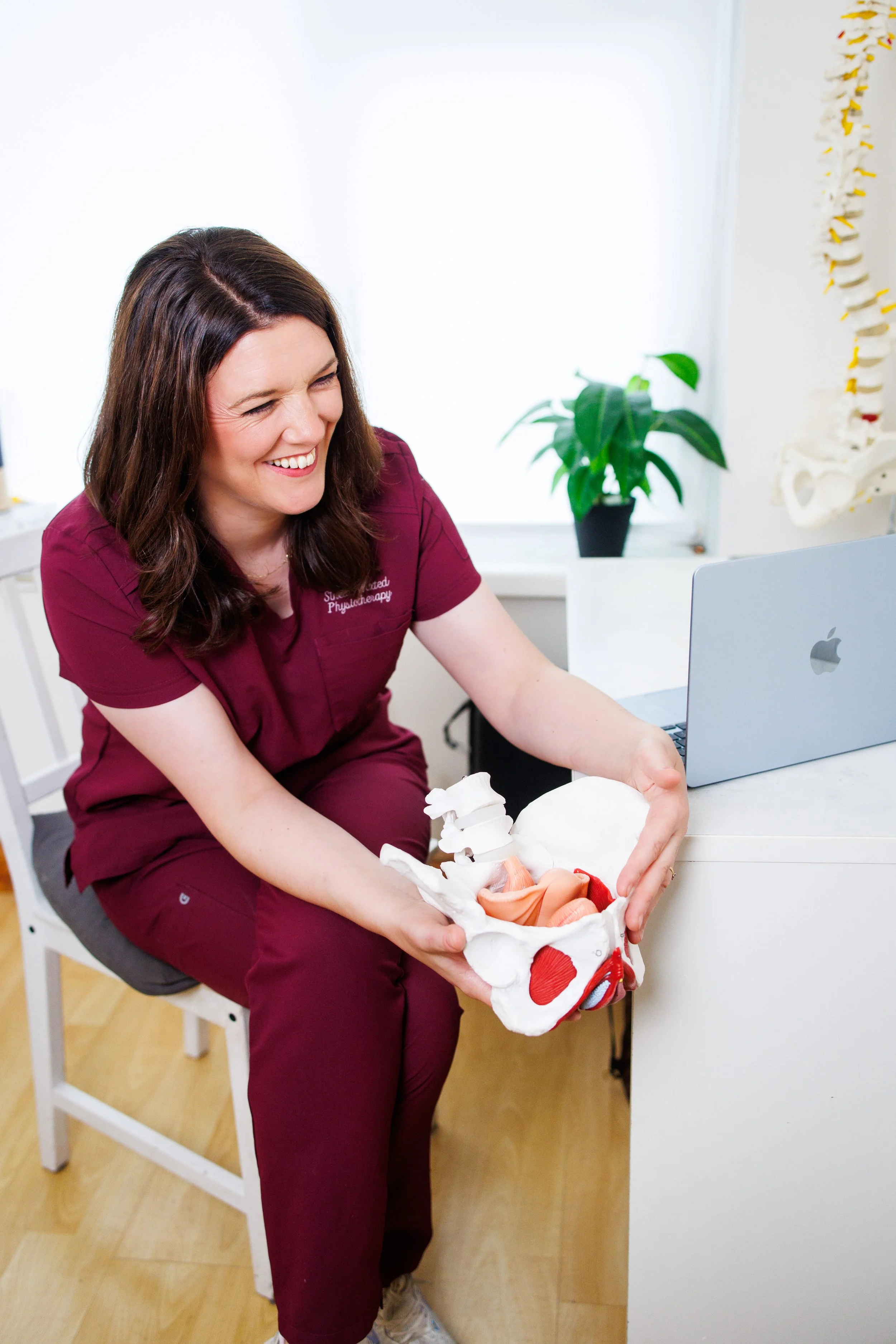 A woman in burgundy medical scrubs sitting on a white chair, smiling and holding a model of a human pelvis and reproductive organs, with a computer and a spine model in the background.