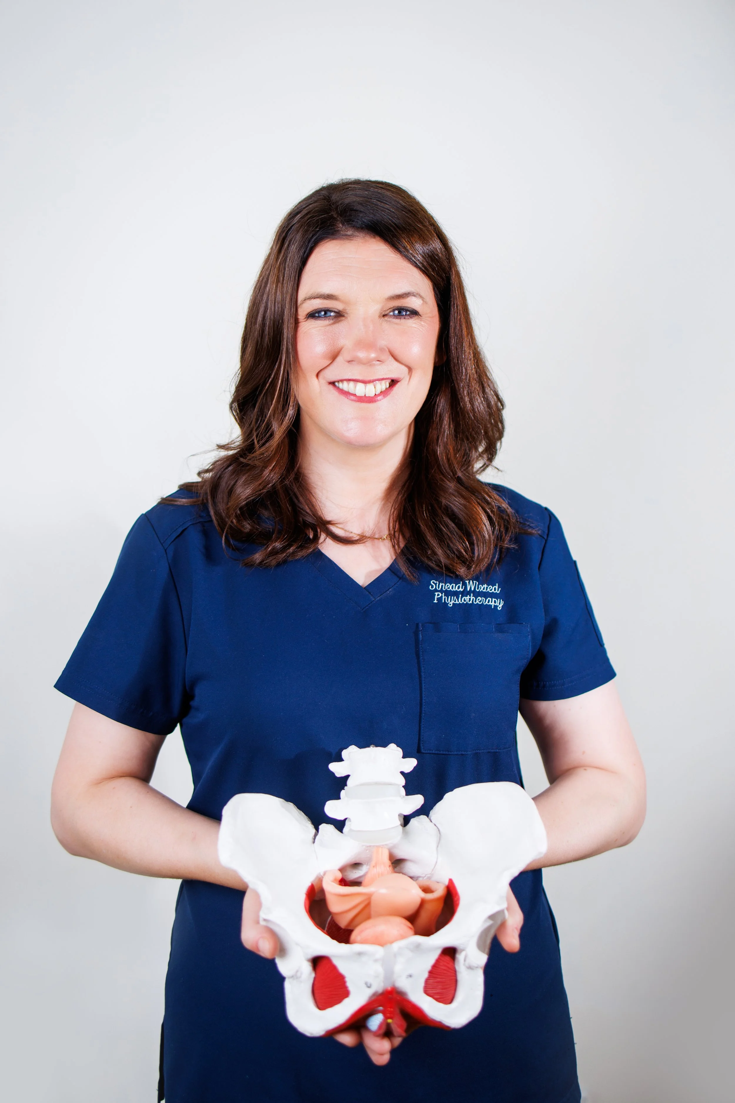 A woman with brown hair and a blue medical uniform holding a model of a human pelvis and lower spine against a plain white background.