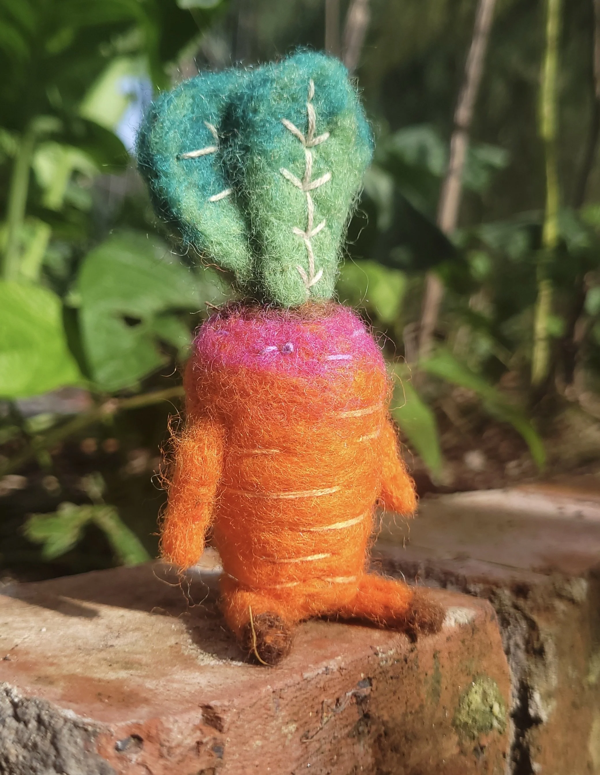Close-up of a small felted carrot with arms and legs, with green, orange and pink fuzzy material, white thread stitched on the green part, and set against a natural outdoor background with green leaves and soil.