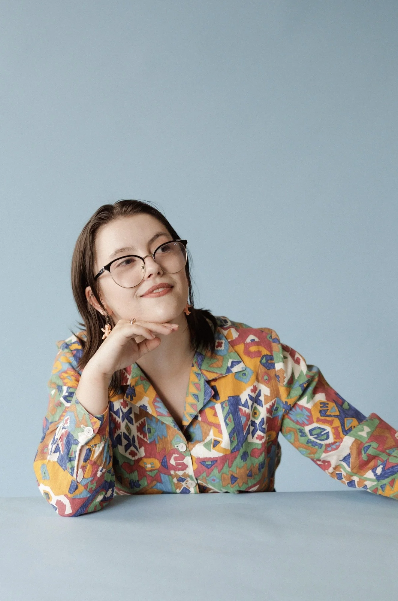 A woman with glasses and shoulder-length dark hair, wearing a colorful patterned shirt, is sitting at a table with her hand resting on her chin against a light blue background.