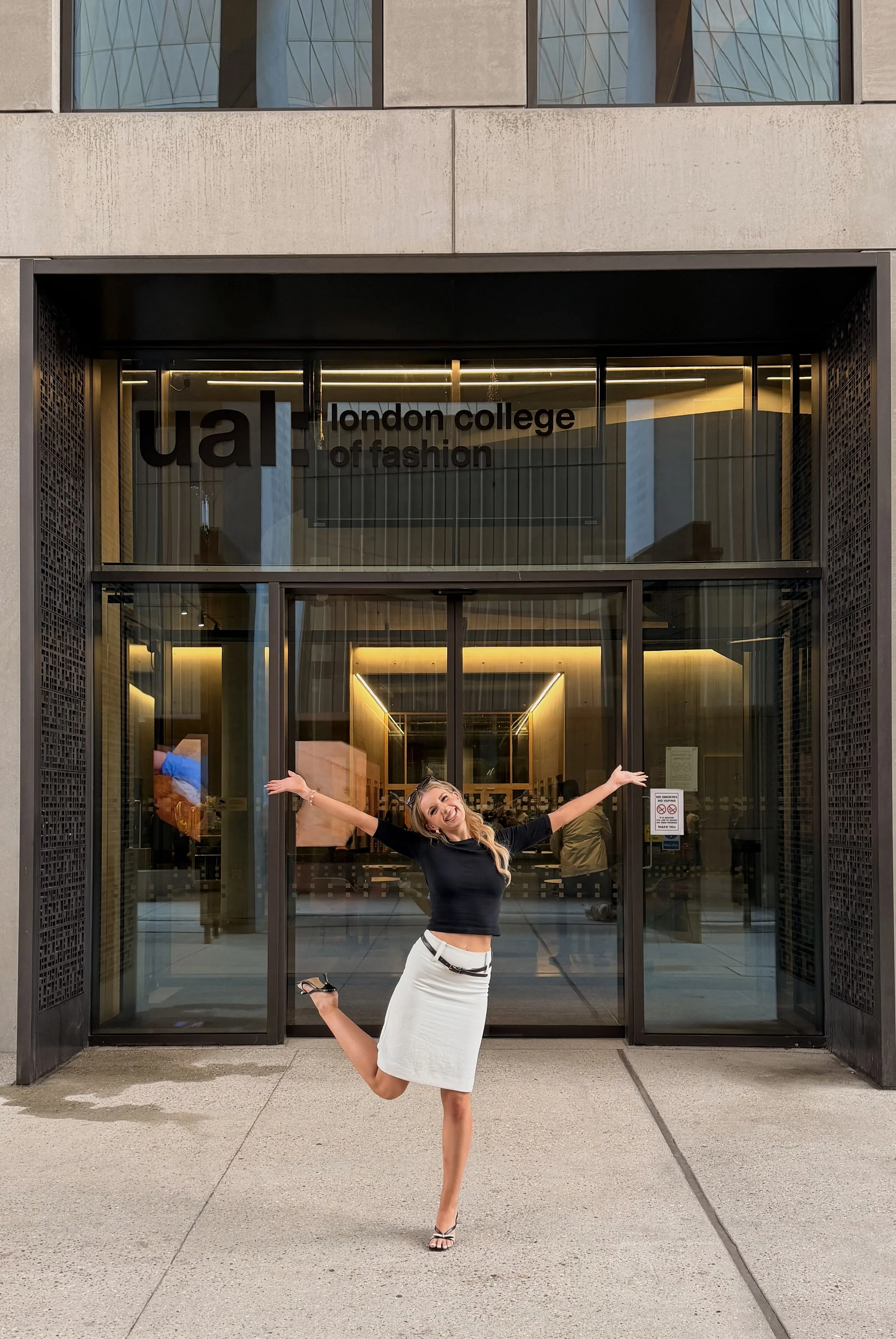 A woman is standing on one leg outside the entrance of London College of Fashion, smiling with her arms outstretched and wearing a black long sleeve top and a white knee-length skirt.