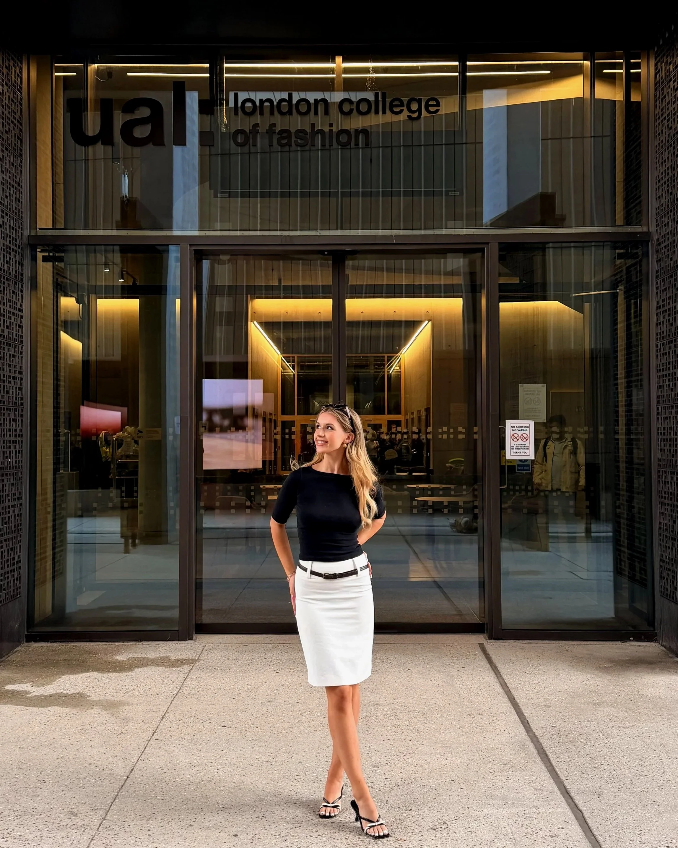 A woman standing outside the entrance of London College of Fashion. She is wearing a black top, white skirt, and black high heels, and is smiling while looking to her left.