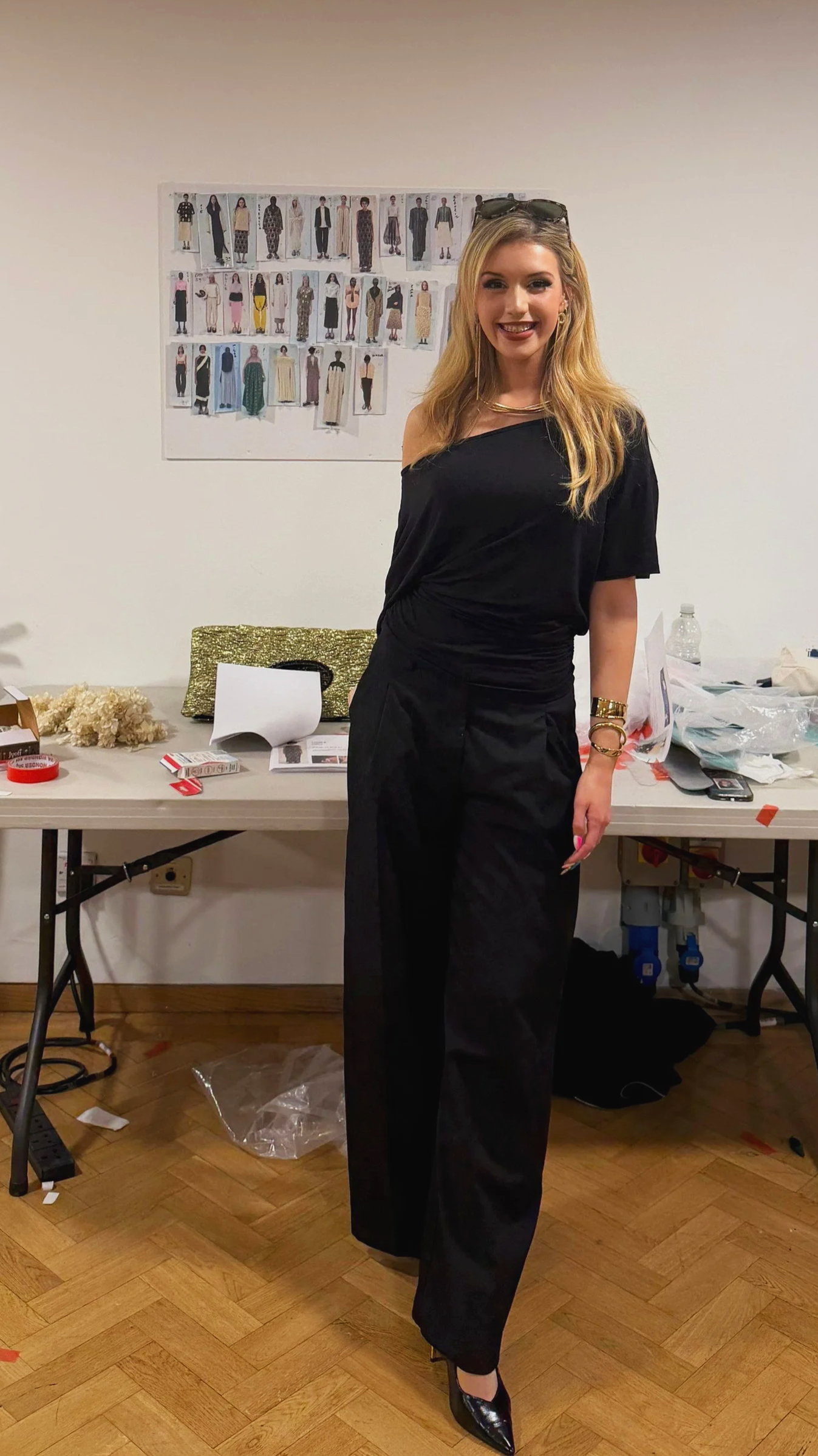 A woman with long blonde hair smiling, wearing a black off-the-shoulder top and black pants, standing in a room with a table and fashion sketches on the wall behind her.