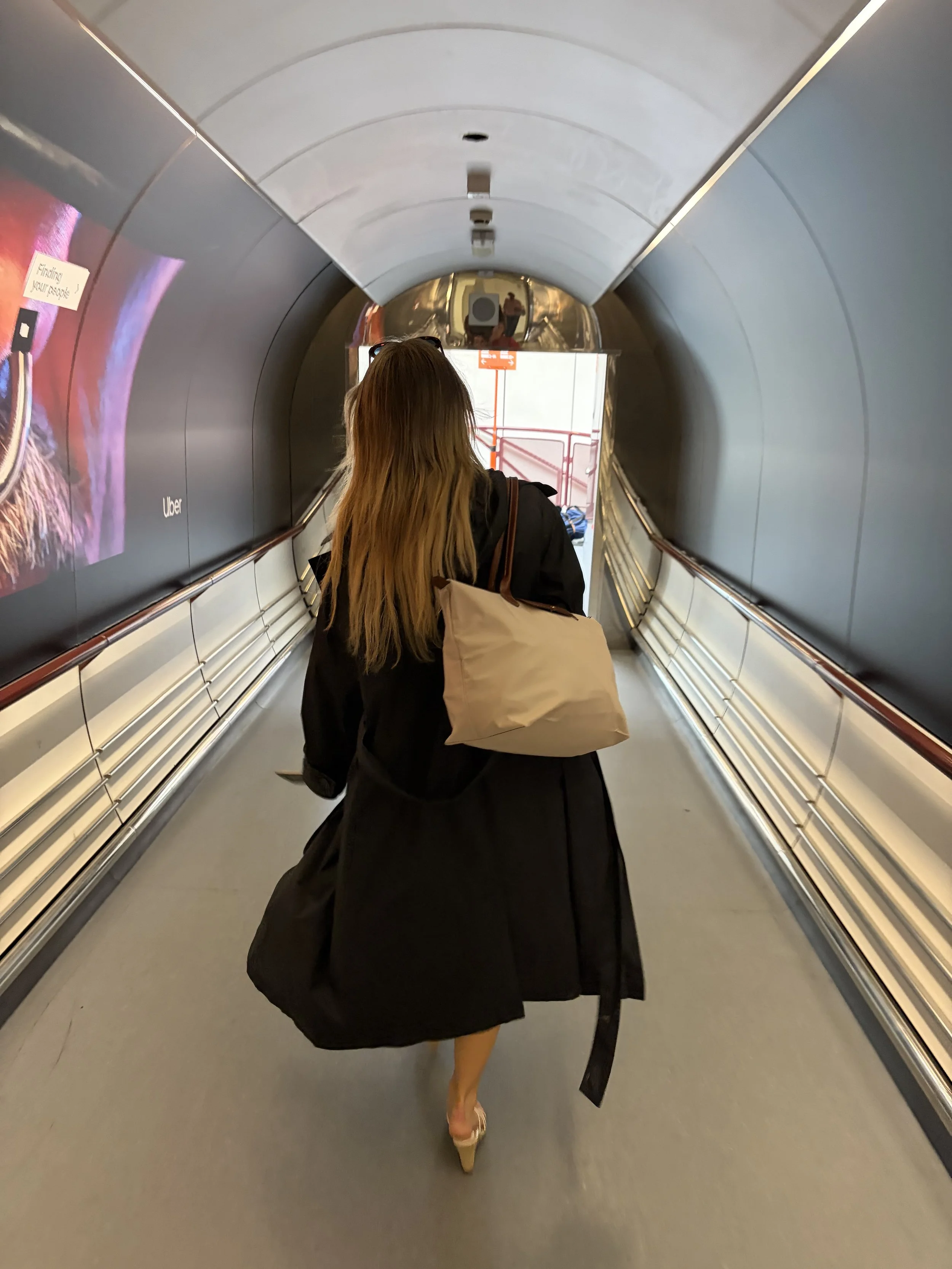 A woman with long hair, walking in a tunnel, wearing high heels and carrying a beige tote bag. She is dressed in a black coat and partial view of her legs.