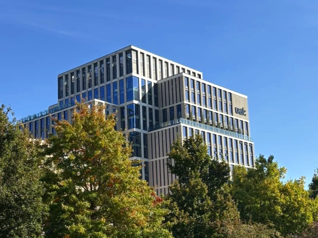 A modern multi-story building with glass windows, partially obscured by trees in the foreground, and a bright blue sky overhead.