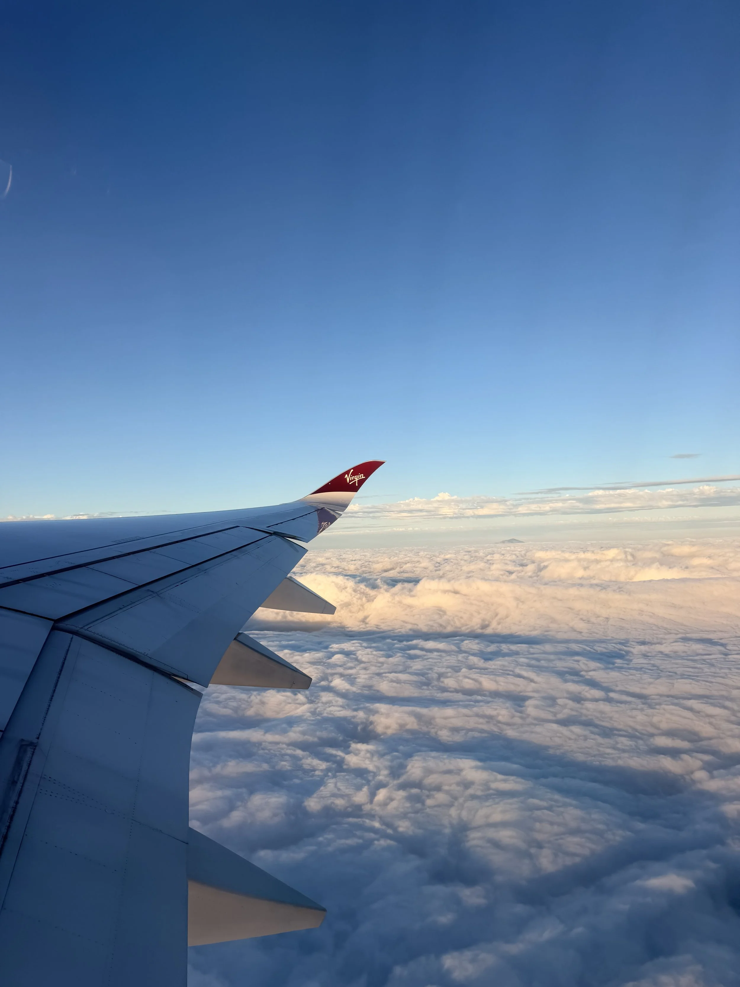 View from airplane window showing aircraft wing with Virgin logo flying above clouds and a clear blue sky.