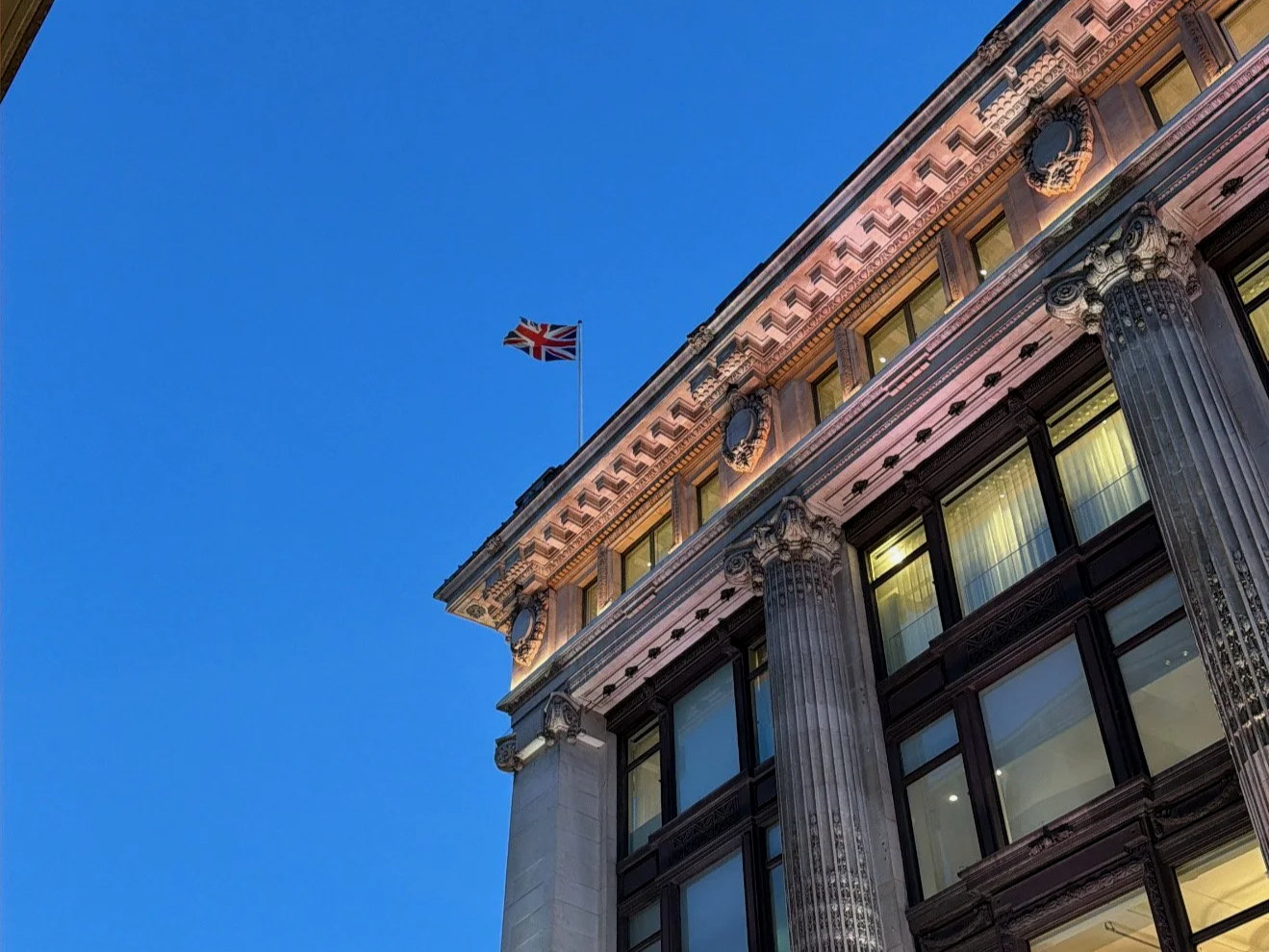 Close-up of a historic building with classical architecture, featuring ornate columns, decorative motifs, and large windows, with a Union Jack flag flying on top against a clear blue sky.