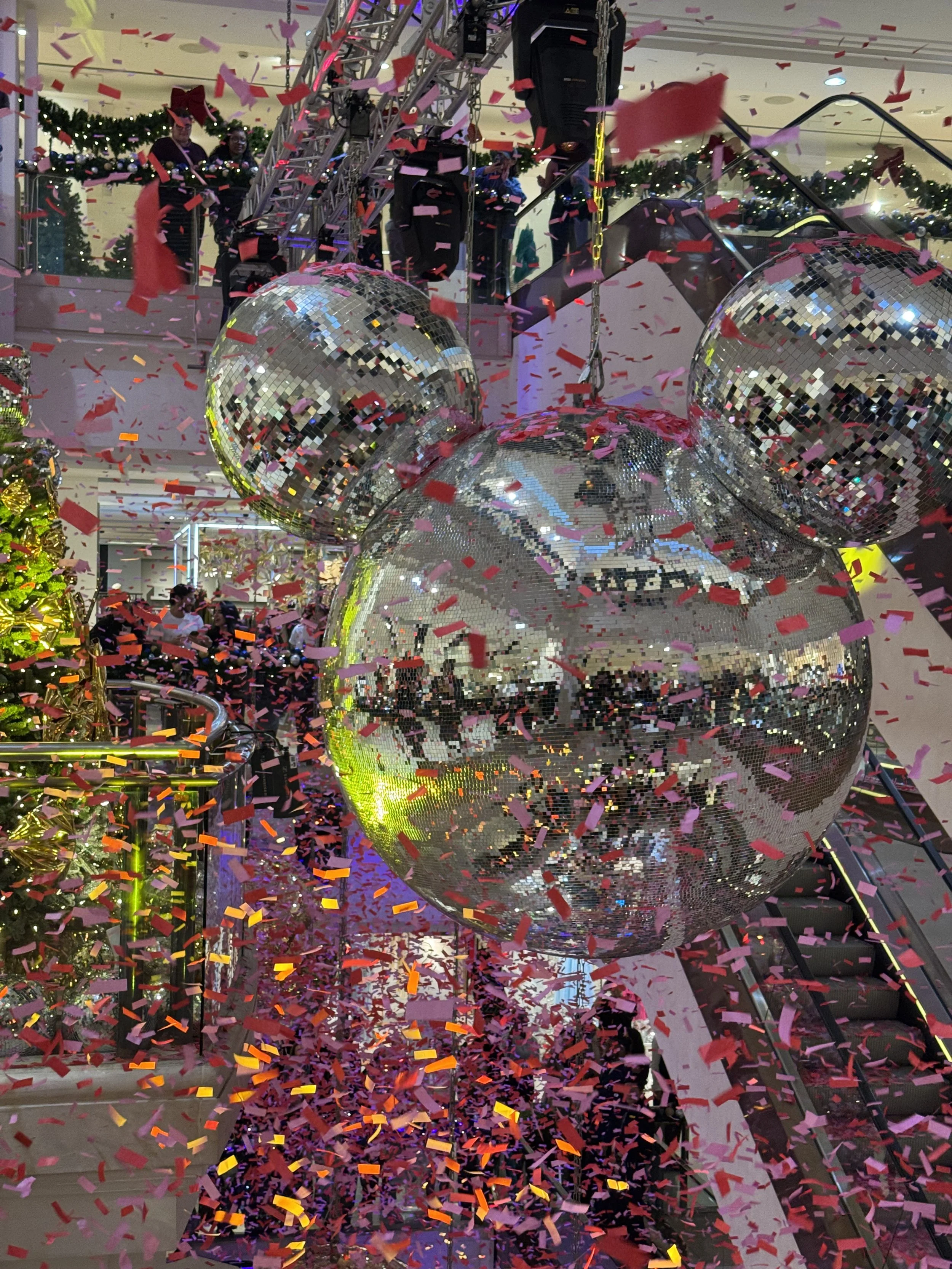 Decorative Christmas display featuring large disco ball ornaments, confetti falling, with Christmas trees and festive garlands in the background, and people observing from the upper level.