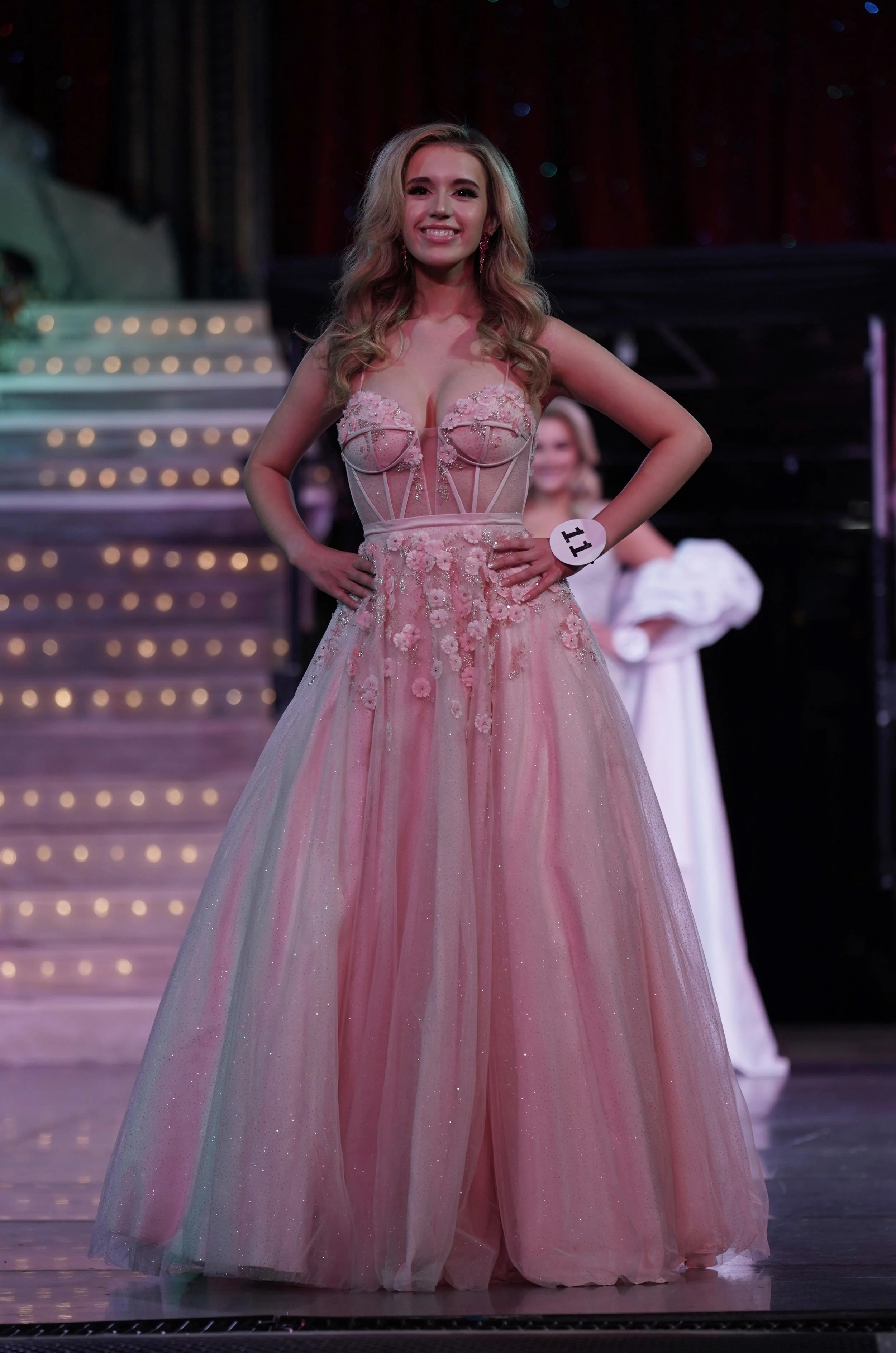 A woman in a pink, floral, ball gown with a deep neckline, standing on a stage with her hands on her hips and a smile. She has long, wavy blonde hair and is wearing earrings. There is a stage behind her with stairs and stage lights, and another woman dressed in white can be seen in the background.