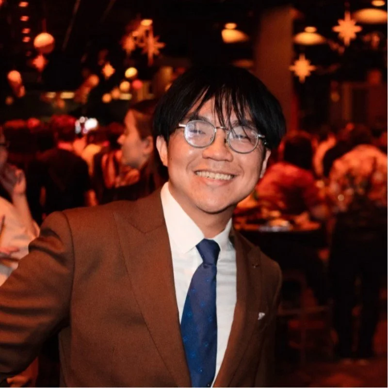 A smiling man in a brown suit, white shirt, and blue tie at a formal event with holiday decorations and other guests in the background.