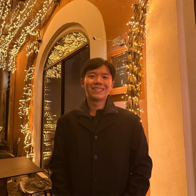 A smiling young man wearing a black jacket stands indoors in a restaurant decorated with Christmas lights and ornaments.