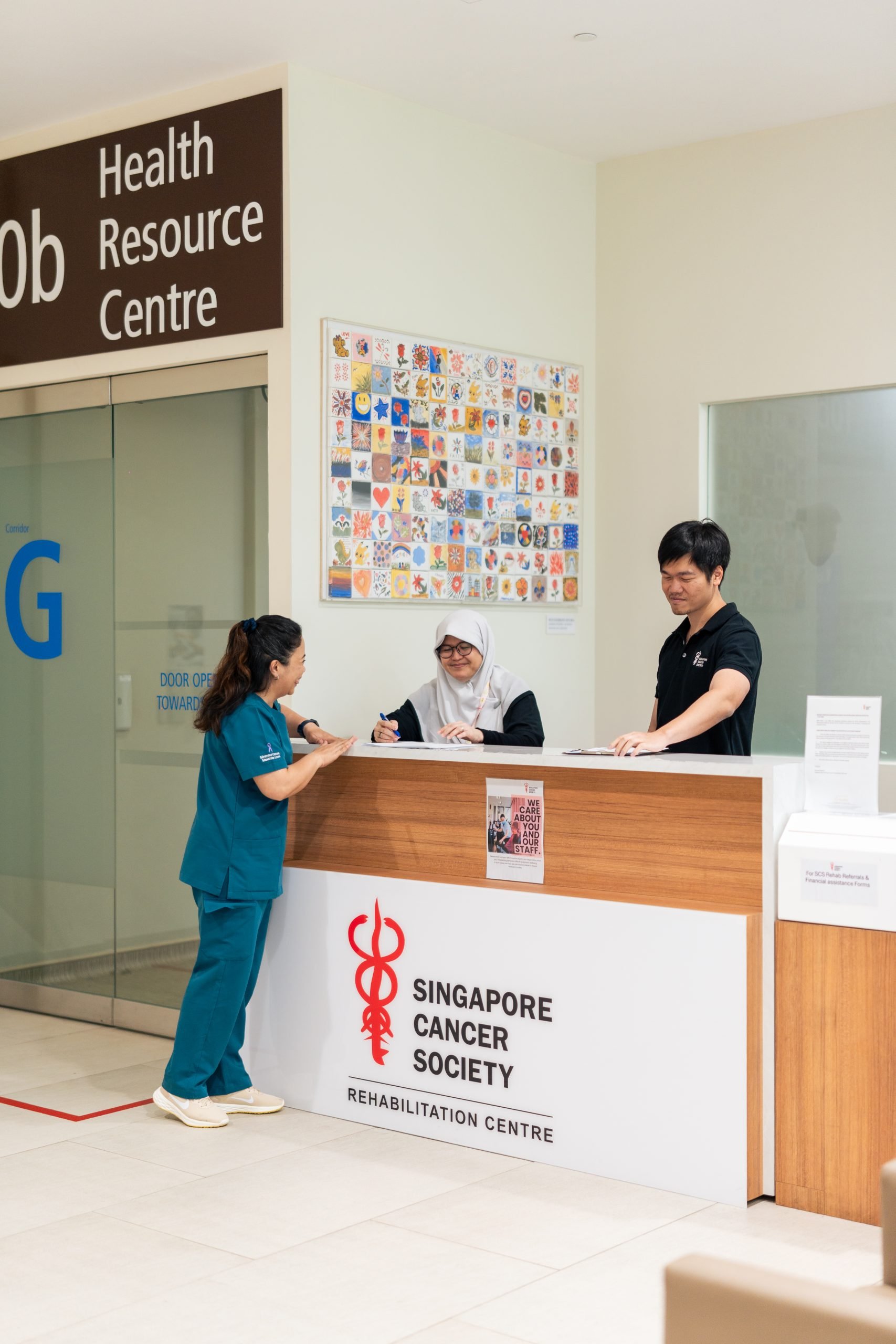 Reception area at the Singapore Cancer Society Rehabilitation Centre, with staff members interacting at the counter, a large sign on the counter, and a colorful piece of wall art behind the desk.