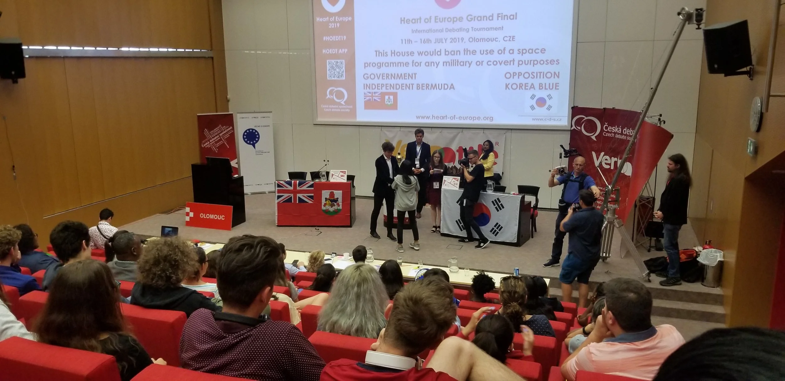 An international debating tournament taking place in an auditorium with people seated and some standing near the stage. Banners and flags from different countries, including the United Kingdom, South Korea, and Bermuda, are displayed. A large screen shows information about the event, including the date and location, and a QR code is visible.