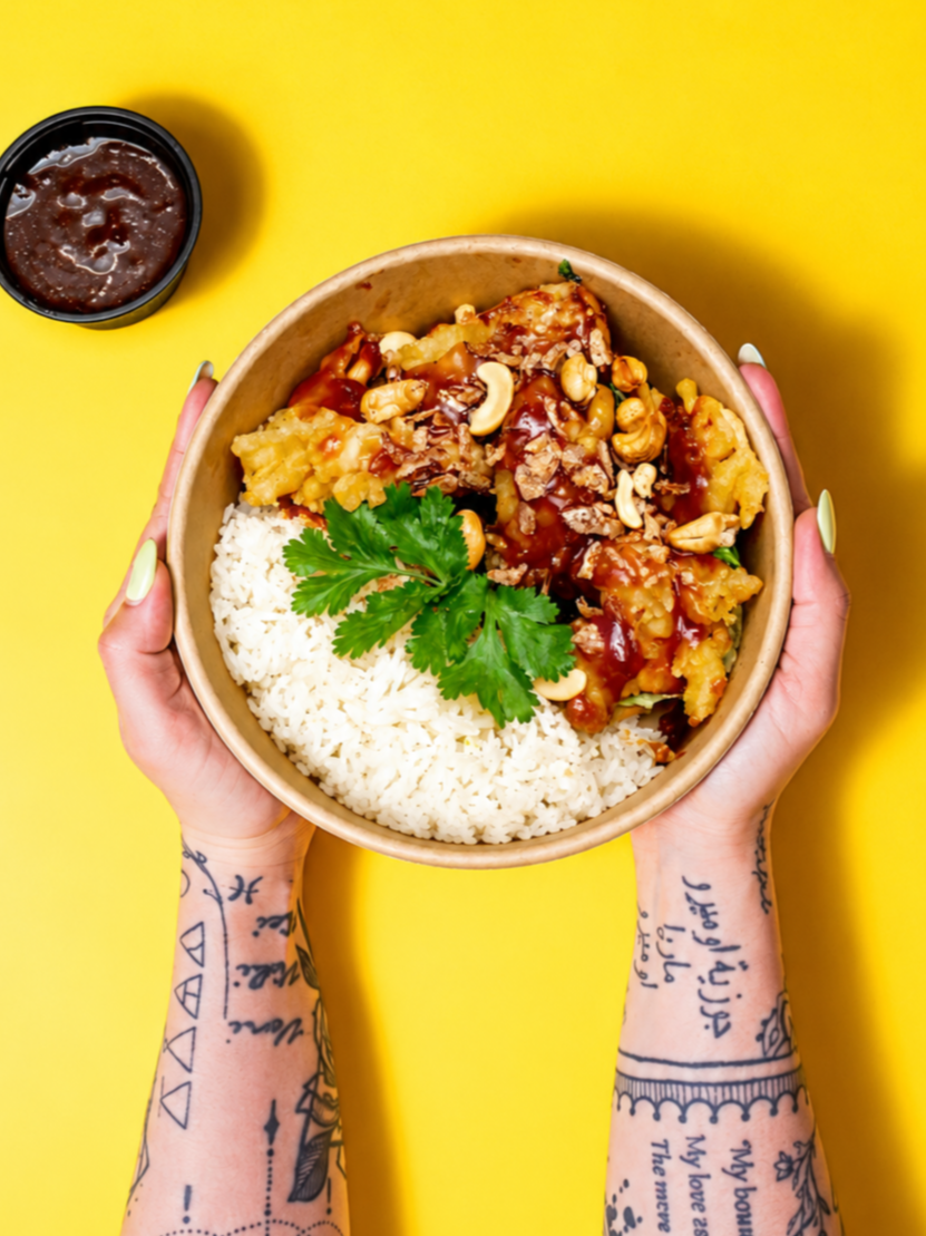 A person with tattooed arms holding a bowl of white rice and chicken with vegetables, topped with chopped nuts and cilantro, on a yellow background. There is a small container of dark sauce nearby.