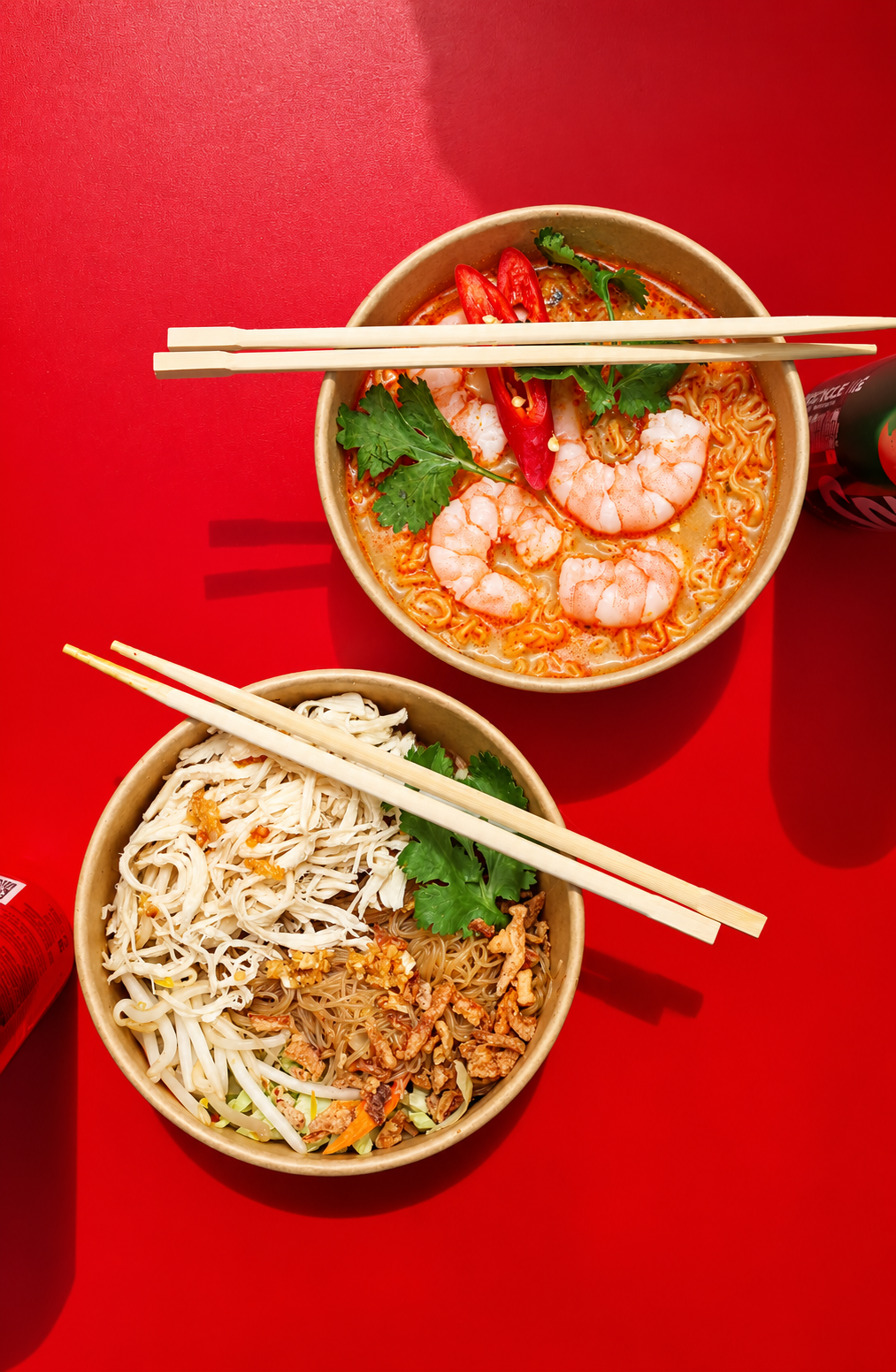 Two bowls of Asian noodle dishes on a red table with chopsticks, garnished with cilantro and chili peppers. One bowl contains shrimp in a spicy broth, and the other has mixed cold noodles with bean sprouts and shredded chicken.