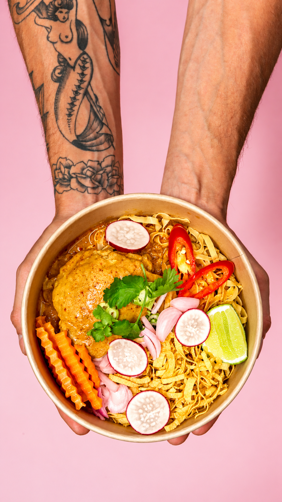 A person with a tattooed arm holding a bowl of Pad Thai noodles with a fried egg, sliced radishes, lime, red chili peppers, carrots, red onions, and fresh cilantro against a pink background.