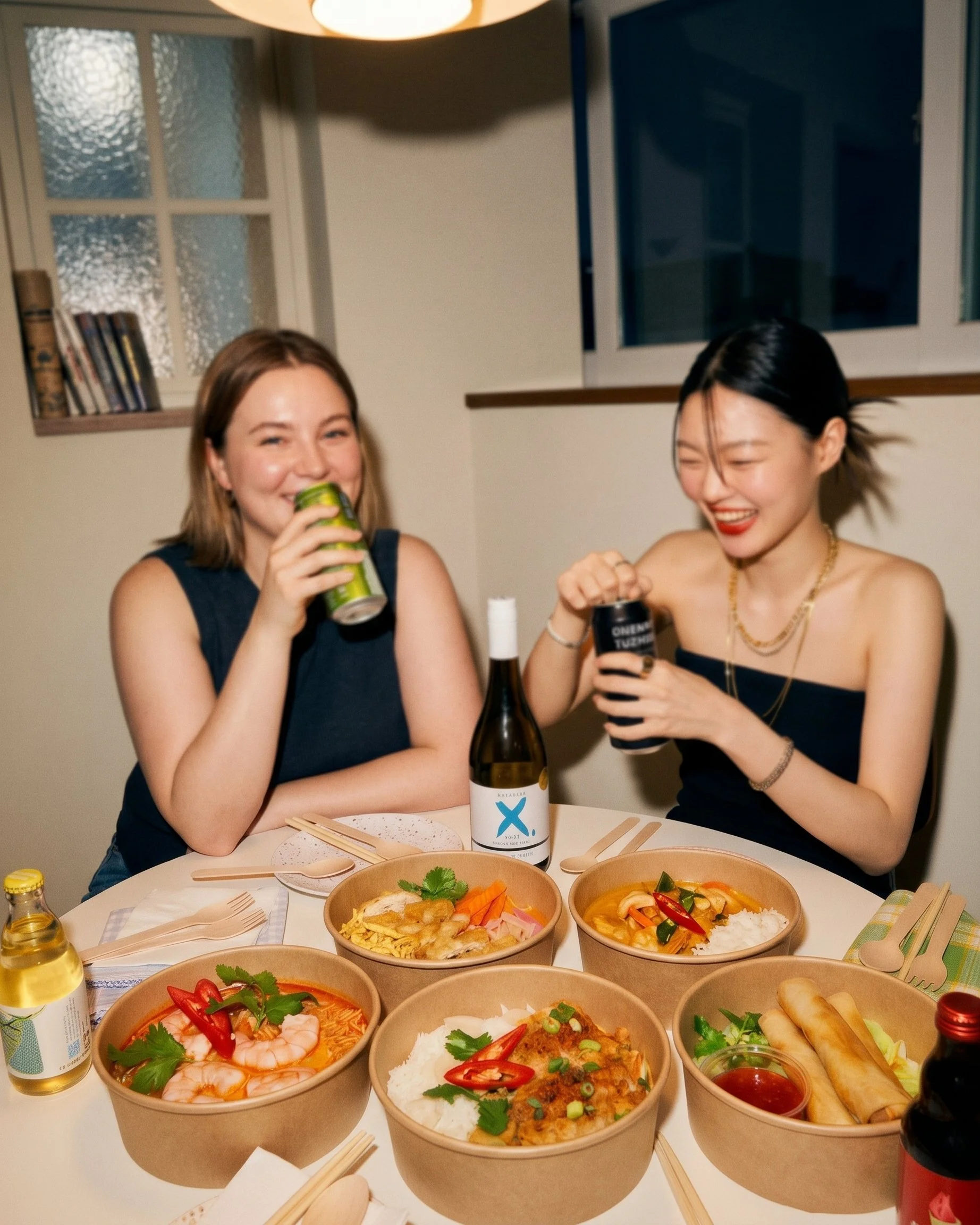 Two women enjoying a meal at a table with several bowls of Asian cuisine, including noodles, rice, and spring rolls, with drinks and condiments.