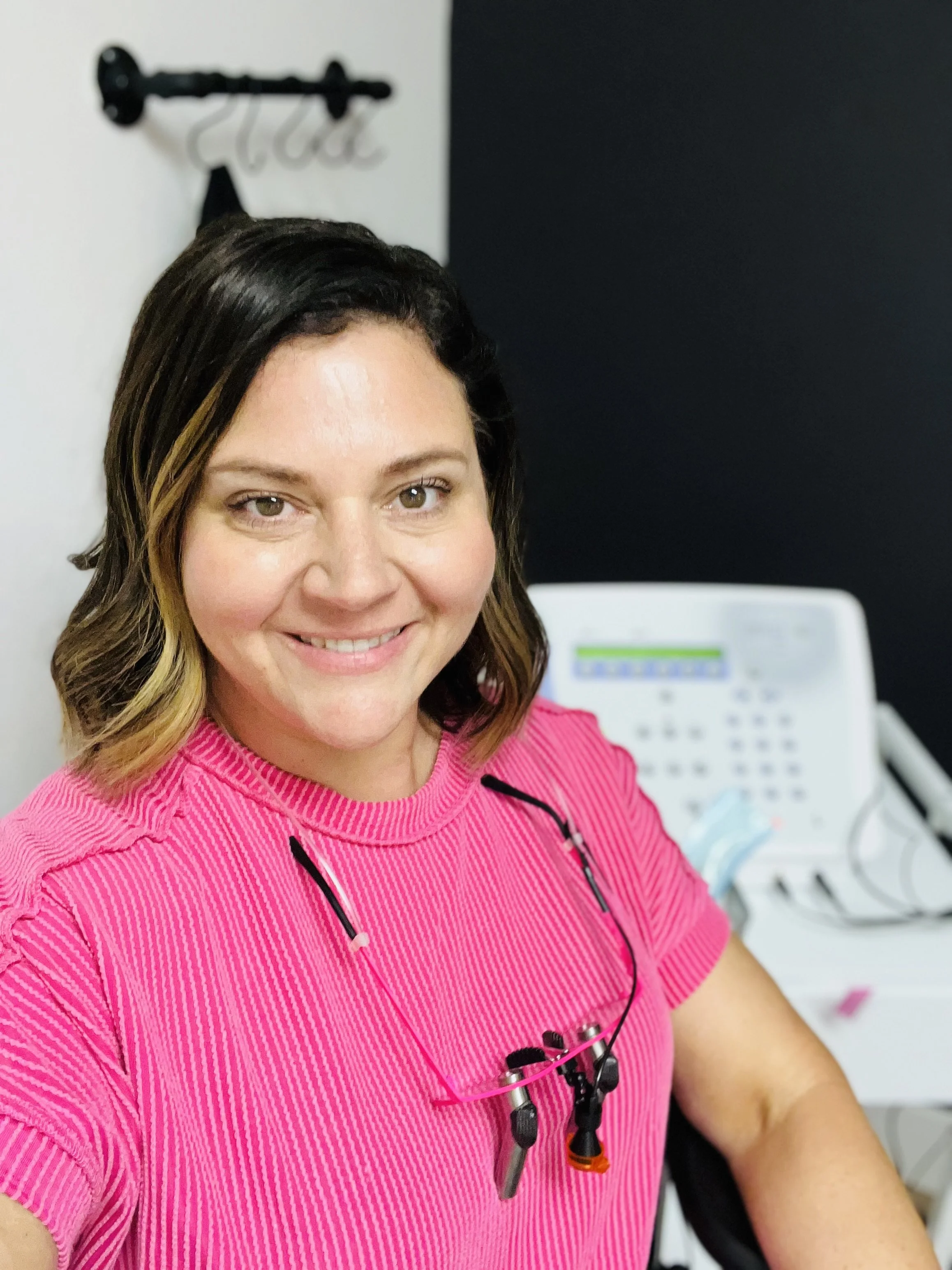 A woman smiling in a pink shirt with headphones around her neck, sitting at a desk with a white telephone in the background.