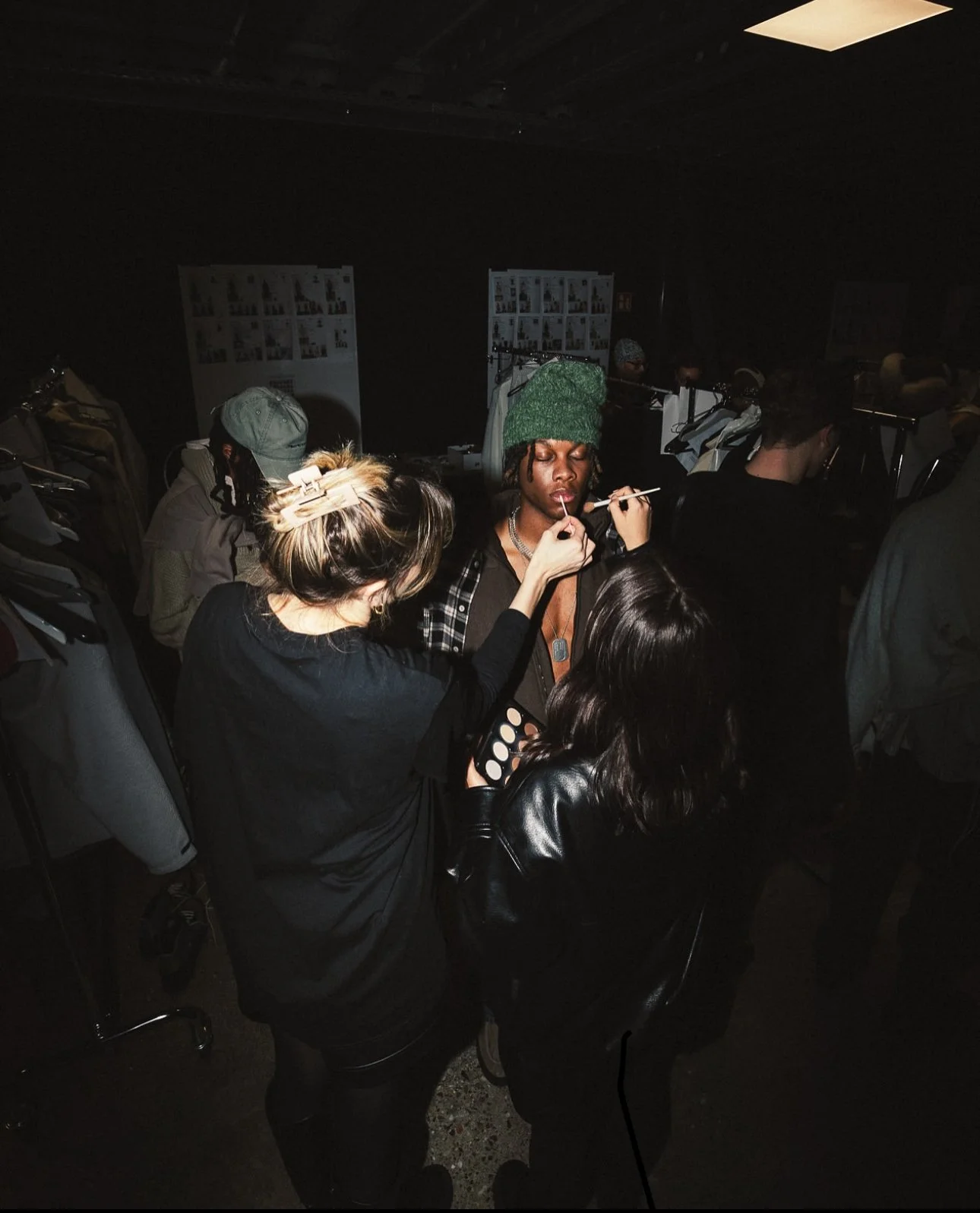A group of people at a backstage makeup area, with a makeup artist applying makeup to a person wearing a green hat, surrounded by racks of clothes and posters on the walls.