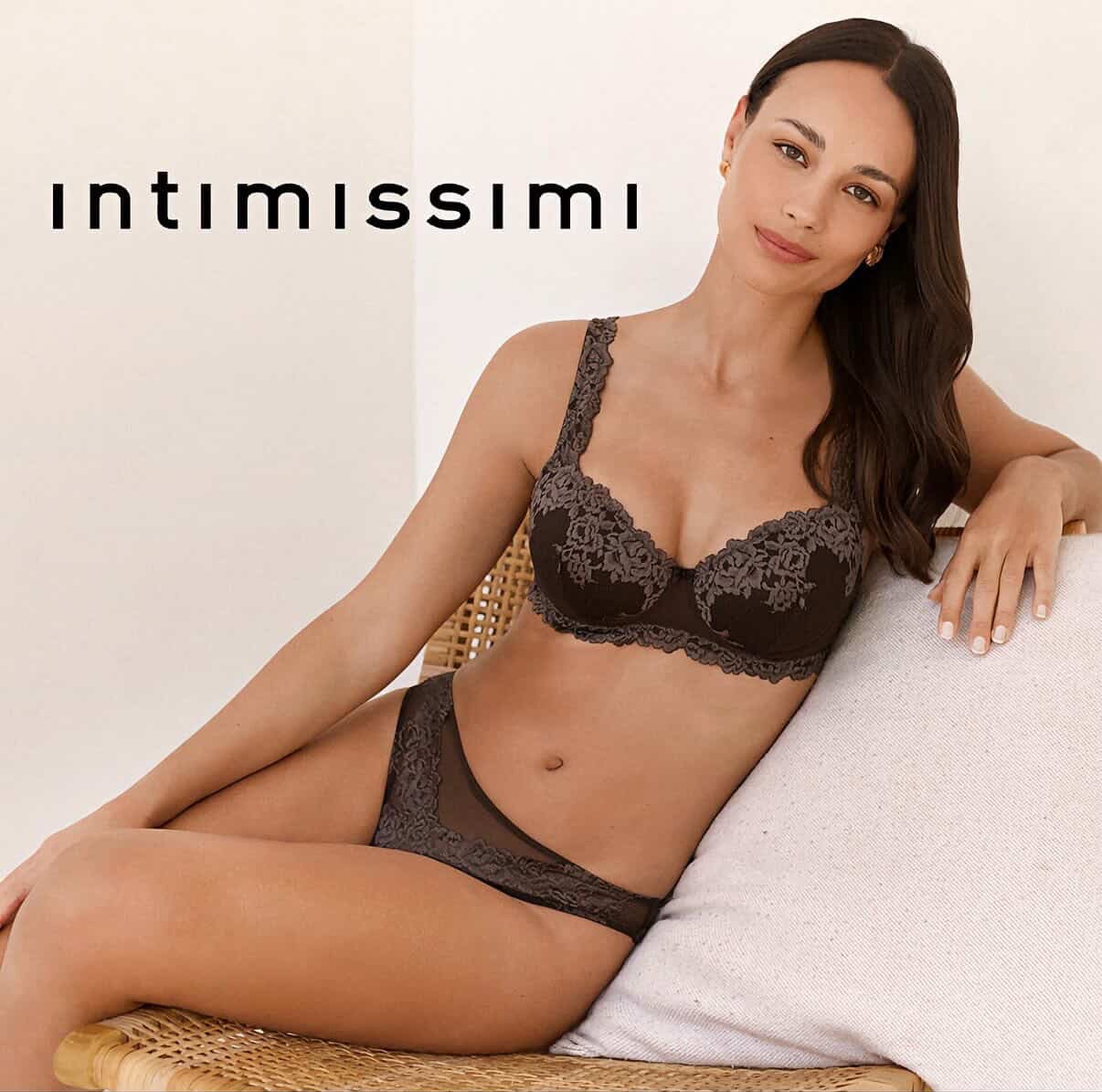 A woman in black lace lingerie sitting on a woven chair with a white pillow, smiling slightly, in front of a beige background with the word 'intimissimi' written.