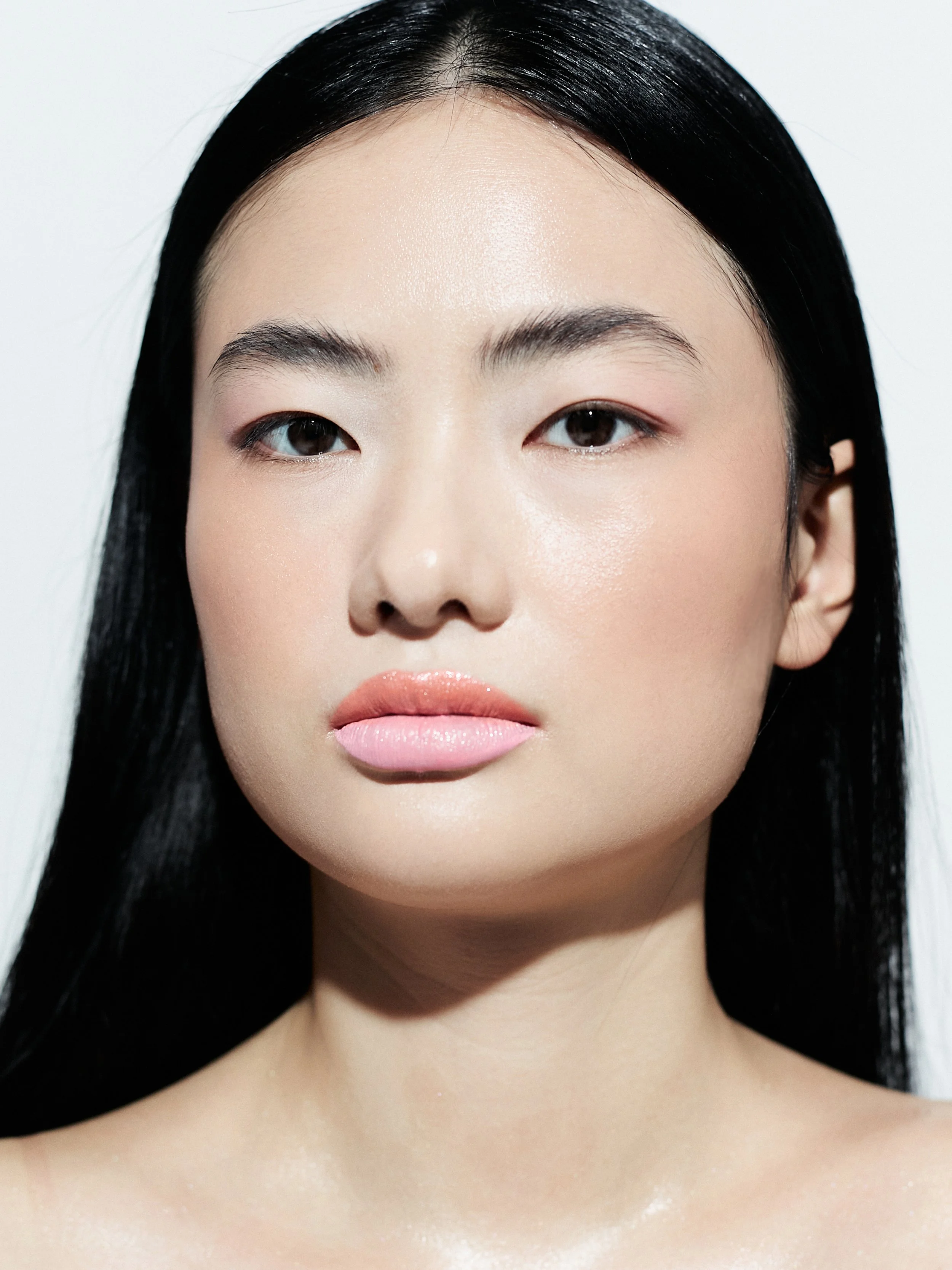 Close-up of a young woman with straight black hair, natural makeup, and light pink lipstick, looking directly at the camera, against a plain white background.