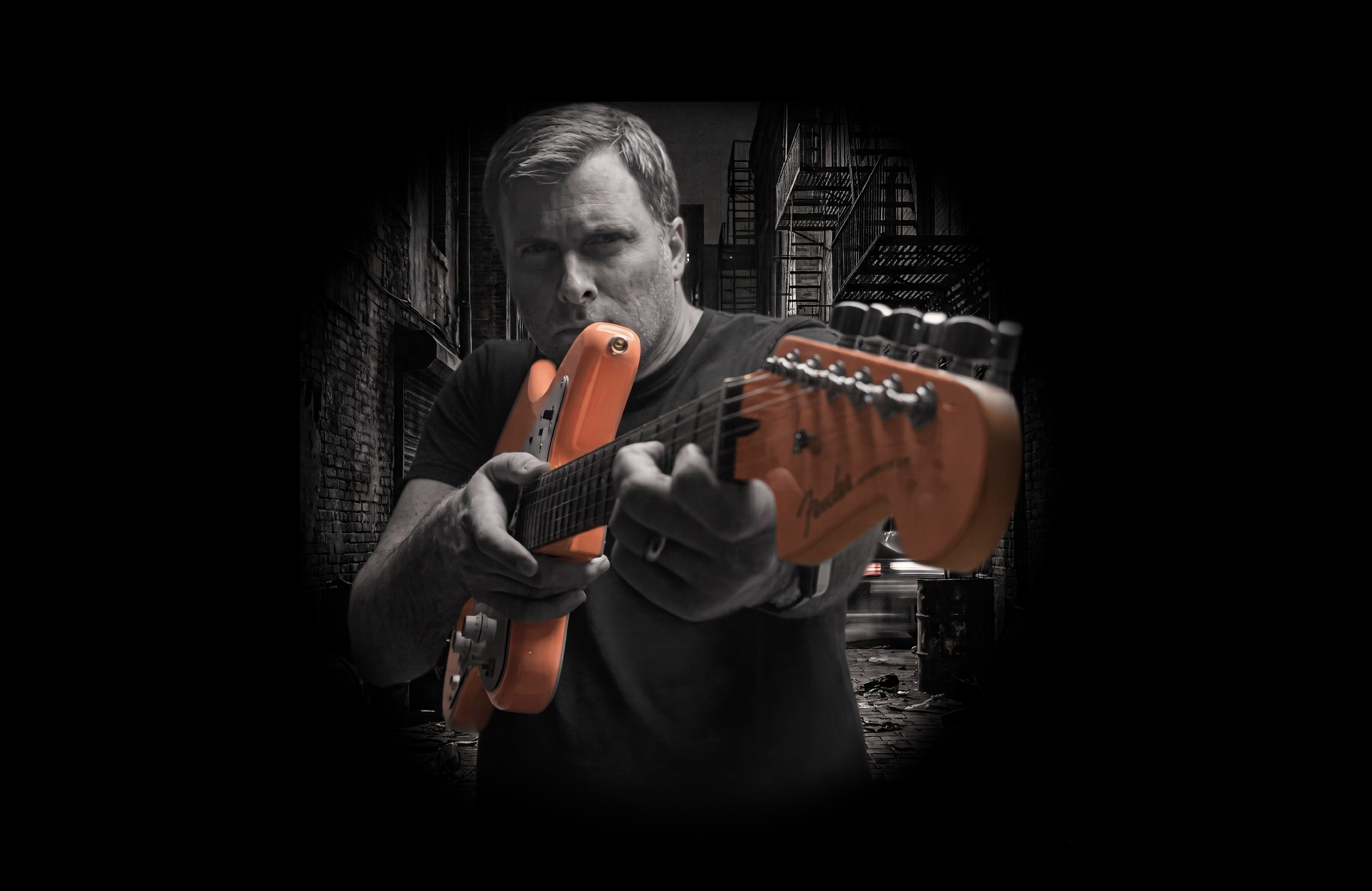 A man with short hair playing an orange electric guitar outdoors in an alleyway at night, with dark brick buildings and fire escapes in the background.