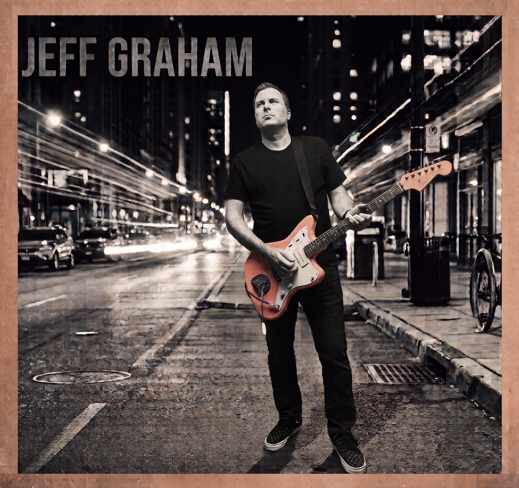 A man is standing on a city street at night, holding an orange electric guitar. The street is wet, with blurred lights from cars and buildings in the background. The man is wearing a black T-shirt, black pants, and black shoes, with a serious expression and looking slightly upward. The scene is illuminated by streetlights and city lights.