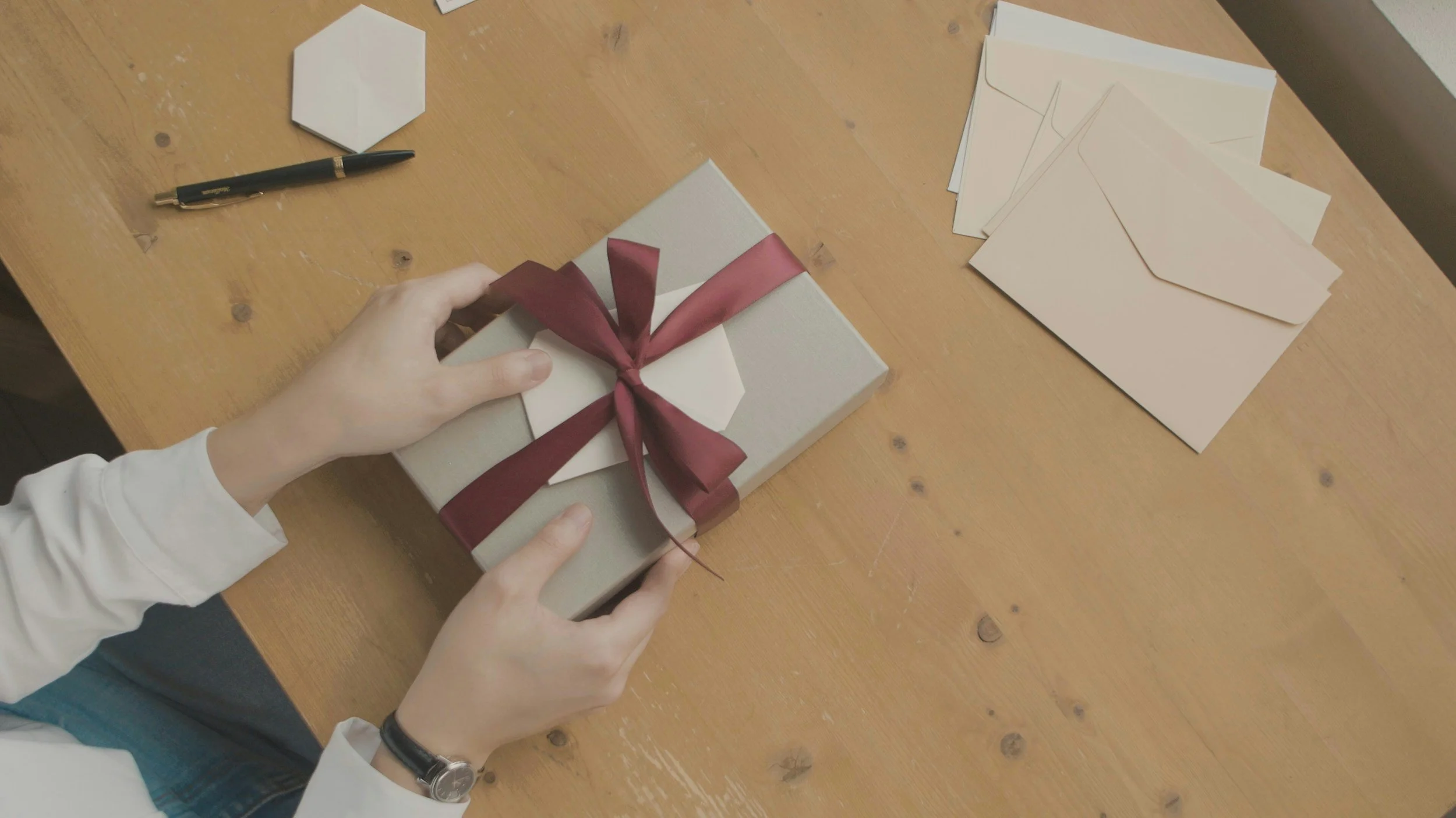 Person holding a wrapped gift box with a red ribbon on a wooden table, surrounded by blank envelopes and a pen.