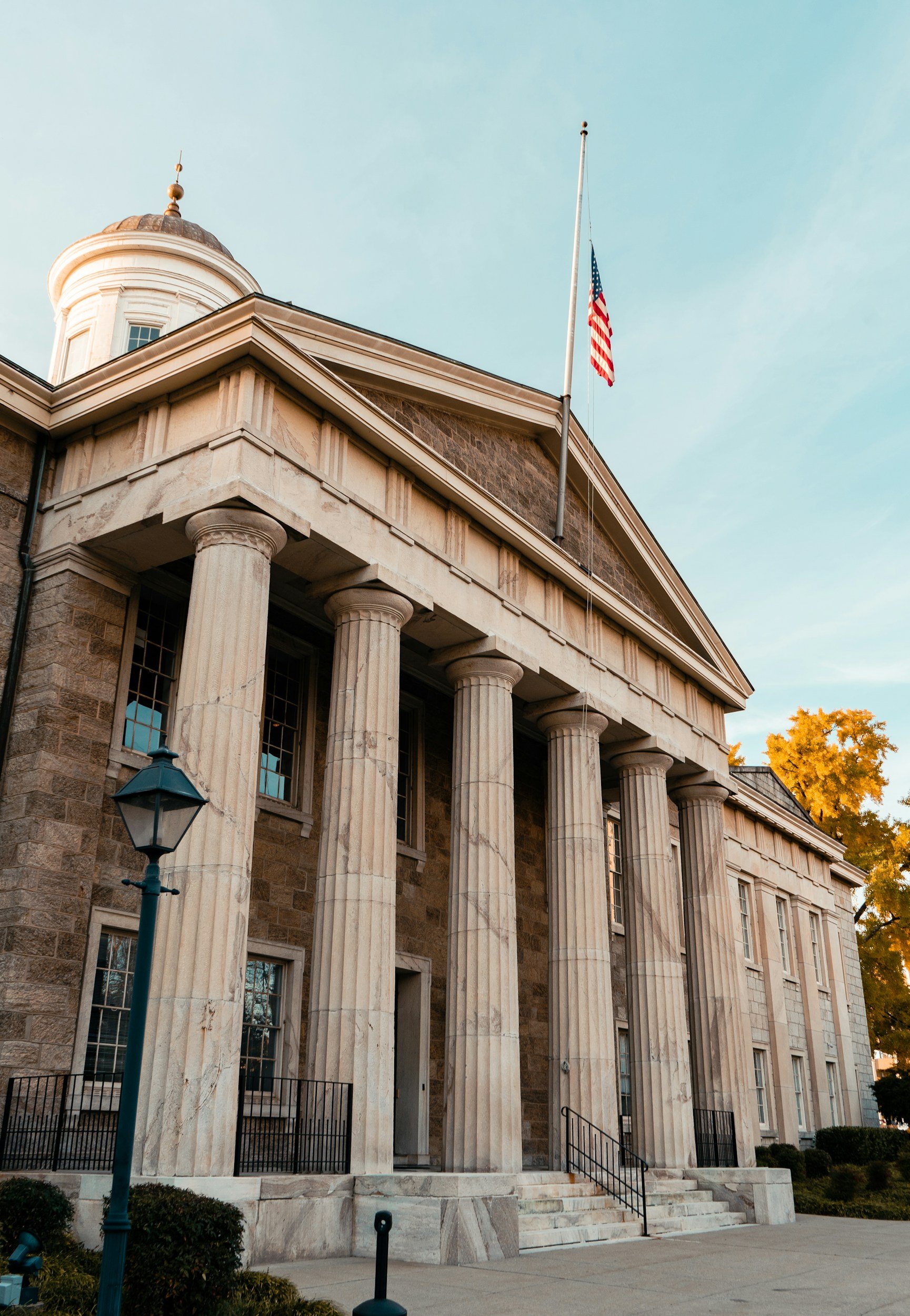 A historic building with tall stone columns and classical architecture, a flagpole with the American flag, and a vintage street lamp in the foreground, during late afternoon with a clear sky and fall-colored trees.