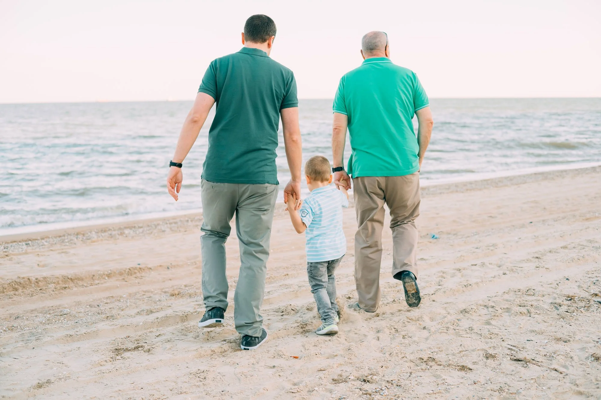 Two men and a boy walking along a beach holding hands, looking at the ocean with a cloudy sky.