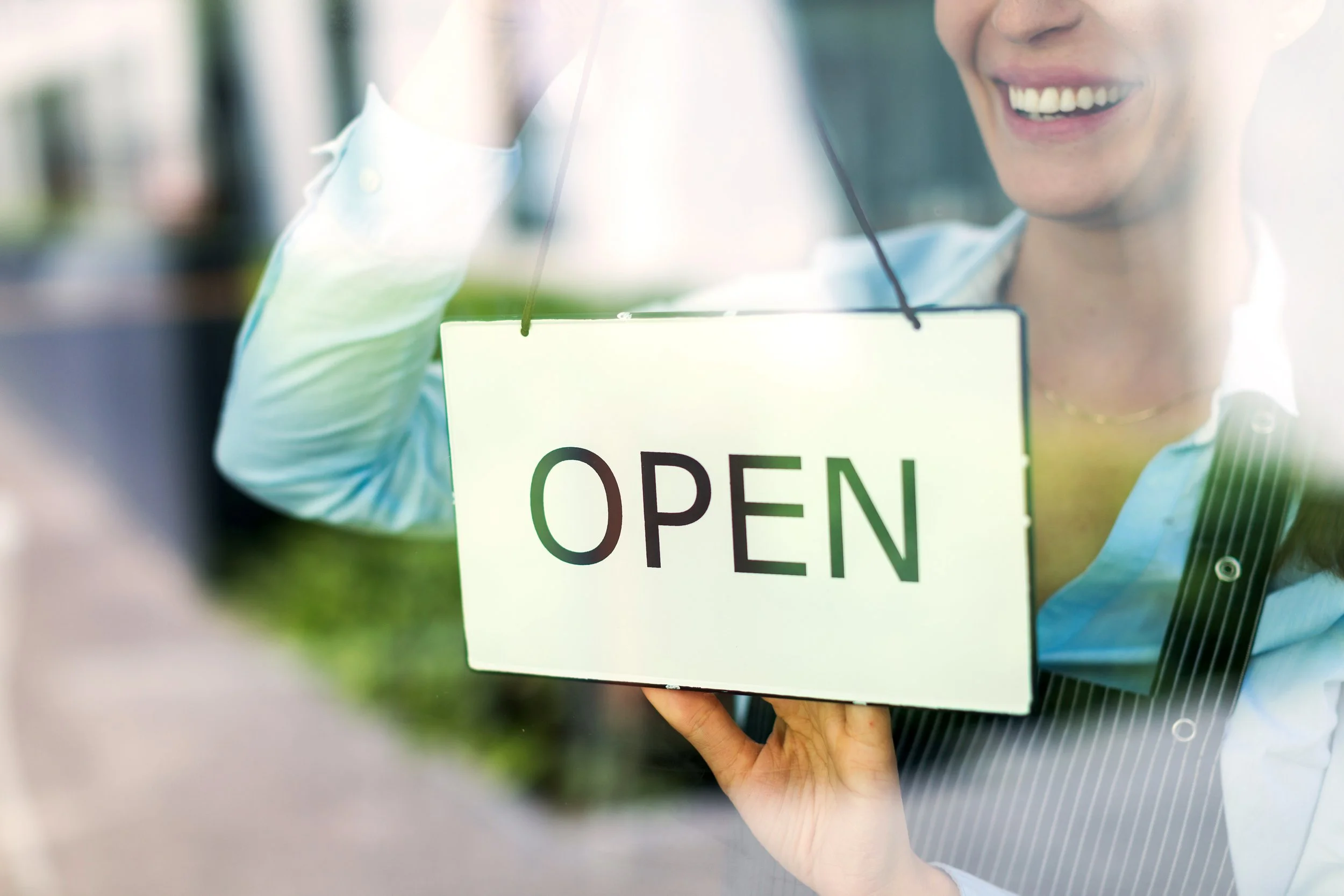Person holding an 'Open' sign on a glass door, smiling.