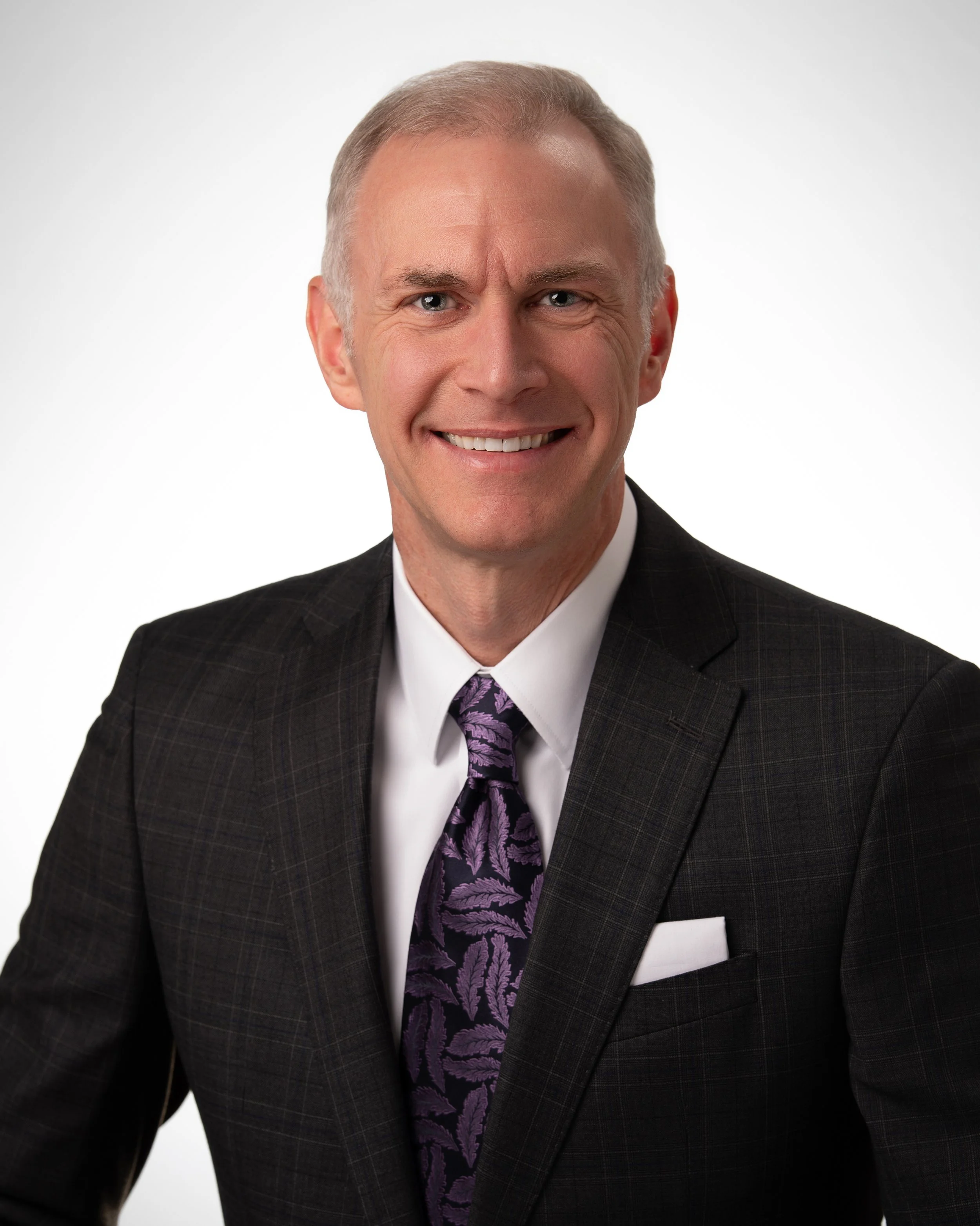 A smiling middle-aged man with short gray hair, wearing a dark plaid suit, a white shirt, and a purple tie with a leaf pattern, against a plain white background.