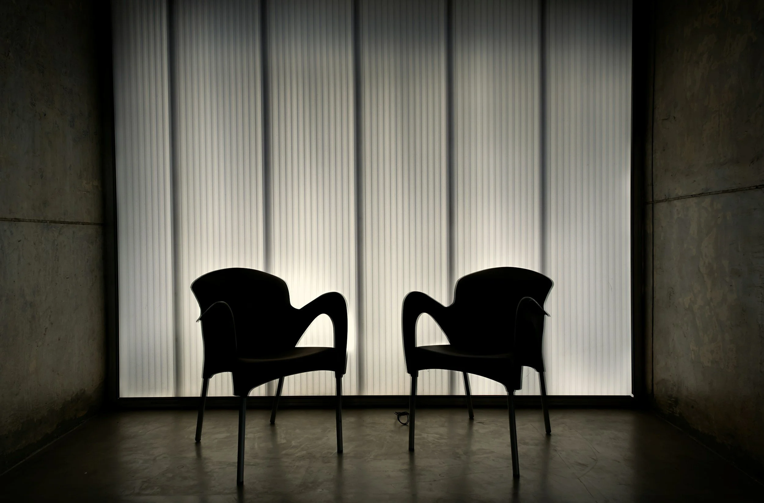 Silhouettes of two chairs in front of a window with vertical blinds, casting shadows in a dimly lit room.