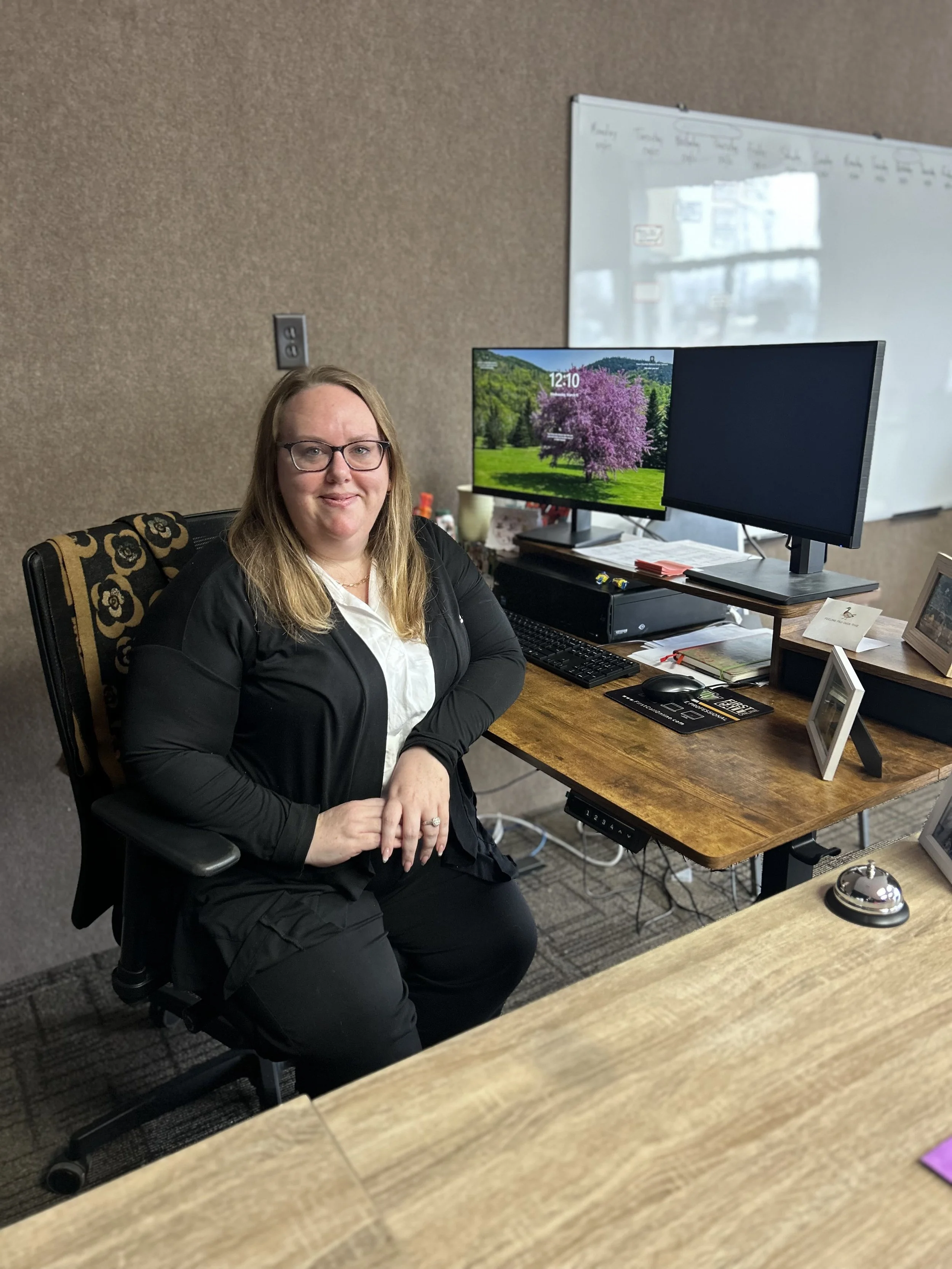 A woman with glasses and long, light brown hair sits at a desk in an office. Behind her are two monitors, one displaying a scenic landscape with a pink flowering tree, and the other turned off. The desk has a black keyboard, a mouse, and various office supplies. There is a whiteboard with writing on the wall behind her, and a beige wall with an electrical outlet.
