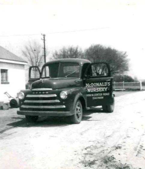 An old black and white truck with a sign for McDonald's Nursery on its door, parked on a dirt road with trees and a house in the background.