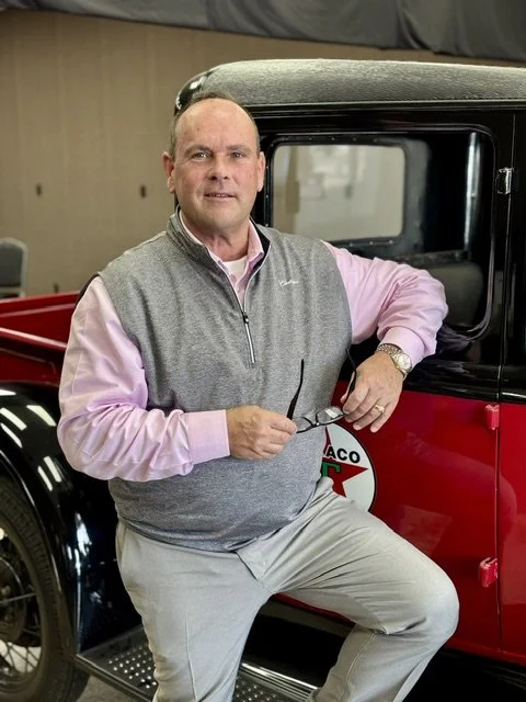 Man standing next to a vintage red truck with a black roof, holding sunglasses, in an indoor garage or showroom.