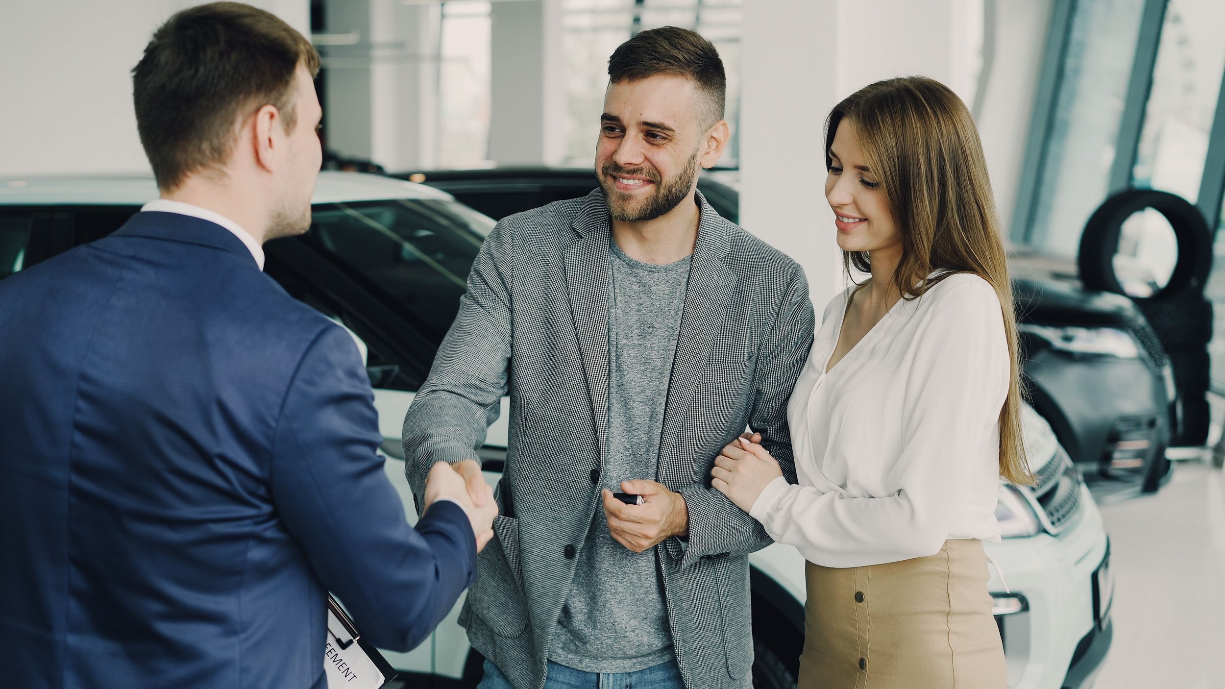 A man in a blue suit shaking hands with a sales representative while a couple looks on happily inside a car dealership.